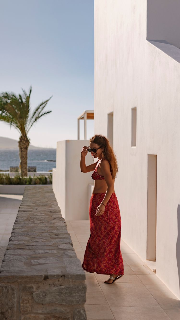 A smiling woman in a red beach outfit walks along a white, minimalist house wall; in the background, the sea.
