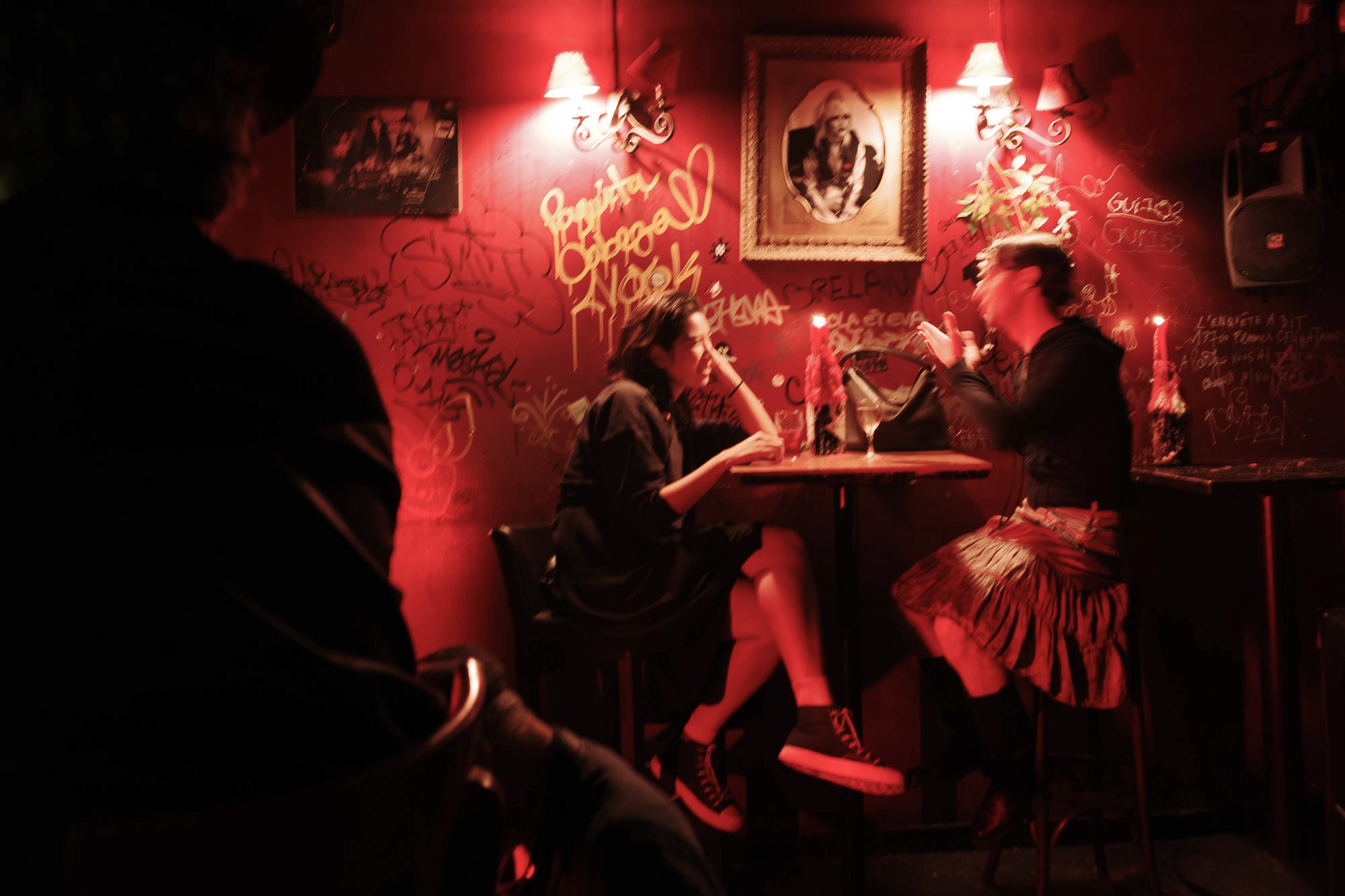 A dimly lit, cozy bar with graffiti-covered red walls, where two people, seated at a round table under warm sconce lighting, engage in animated conversation over drinks, surrounded by eclectic decor.