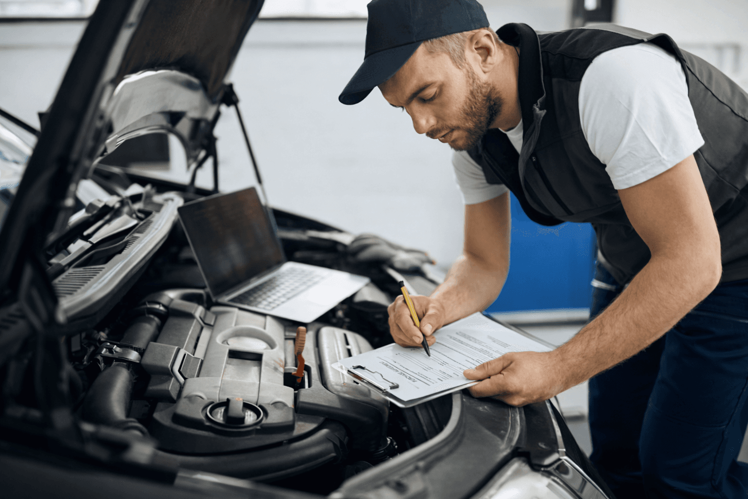Mechanic inspecting a car engine and taking notes beside a laptop.