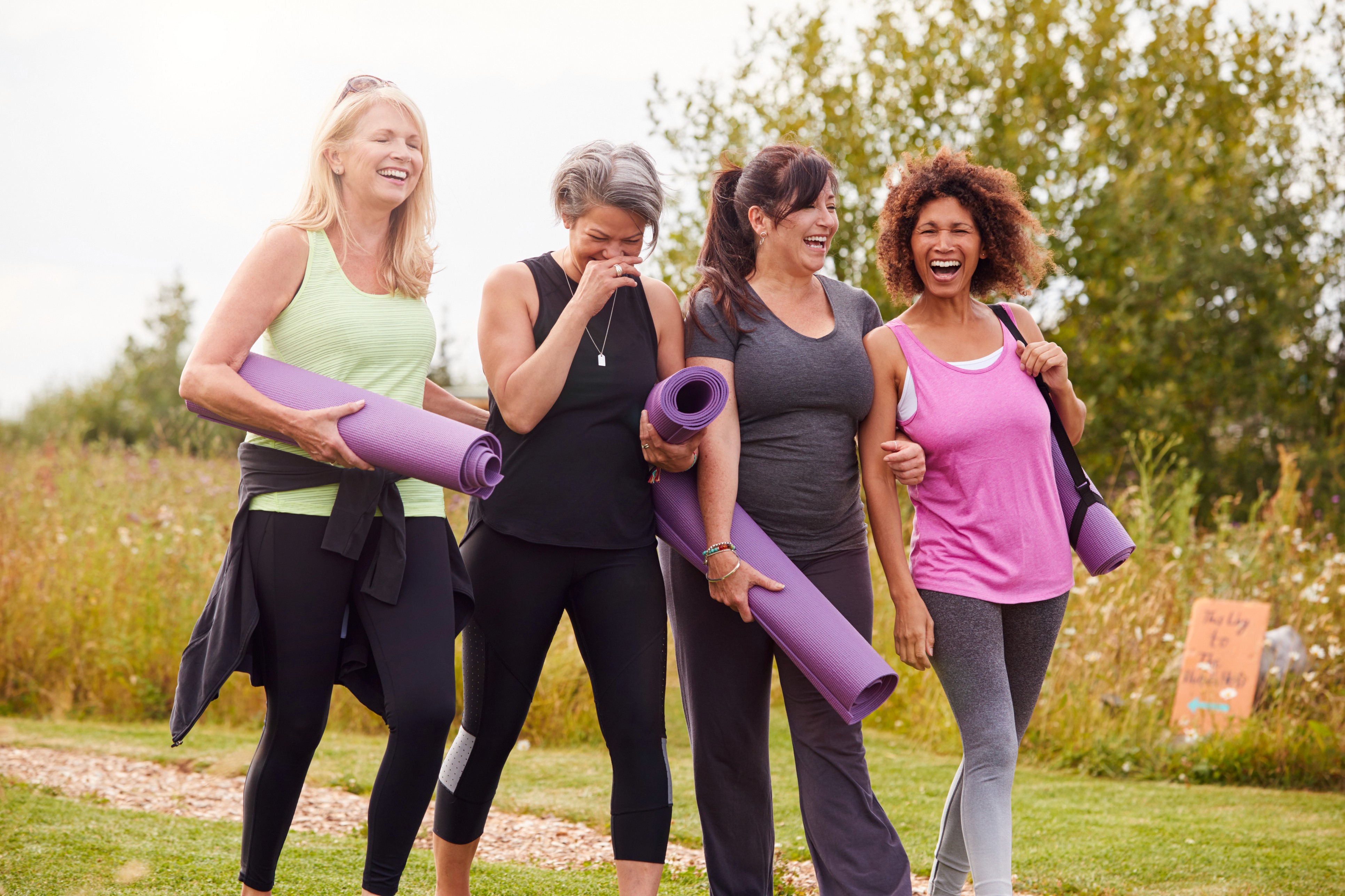 Das ist ein Bild auf dem 4 sportlich gekleidete Frauen in unterschiedlichem Alter zu sehen sind. Sie haben fröhliche Gesichter und halten Fitness Matten in ihren Händen. Im Hintergrund ist die grüne Natur zu sehen.