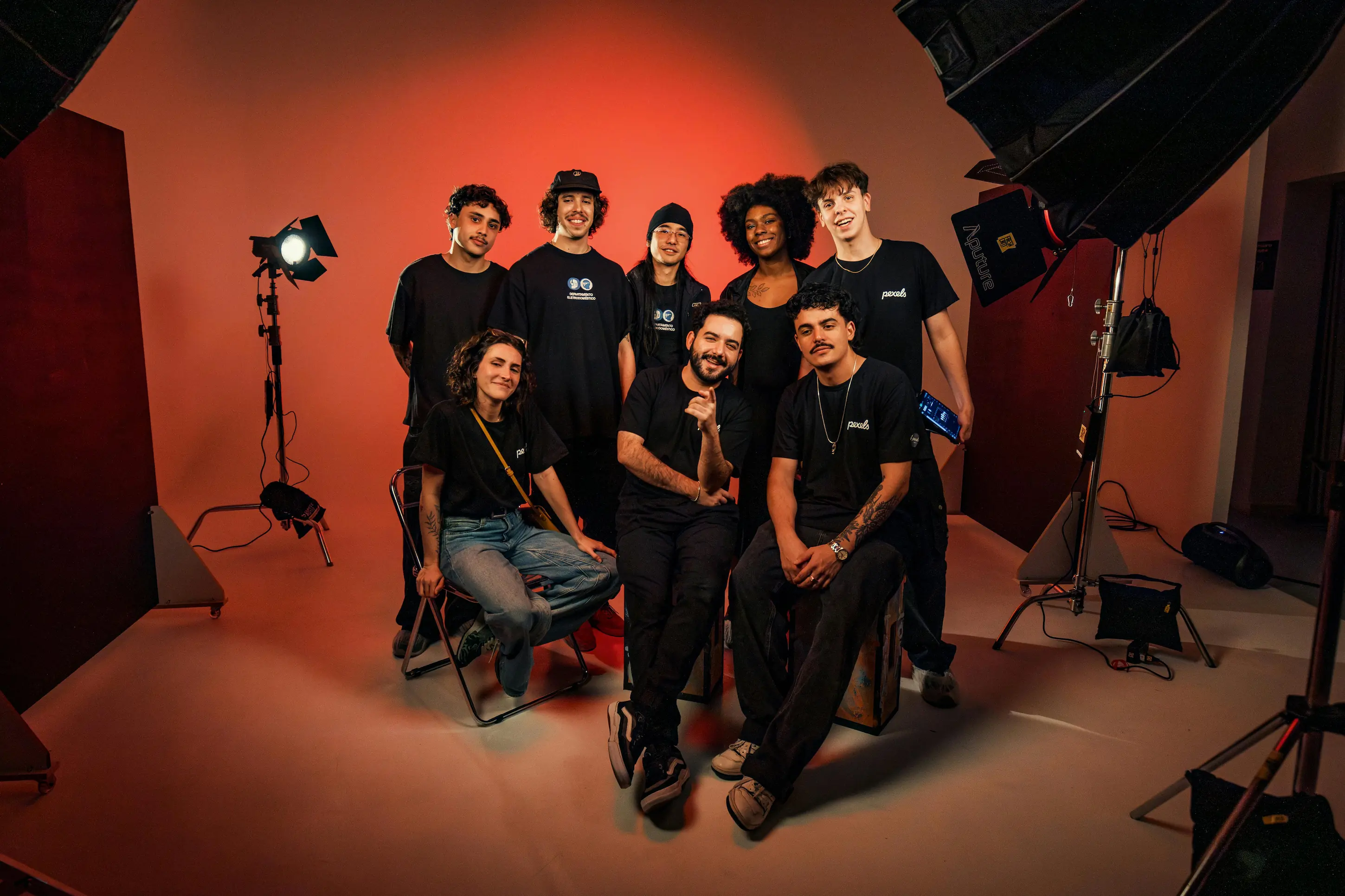 Eight diverse, smiling people pose in a photography studio with professional lighting and a red backdrop.
