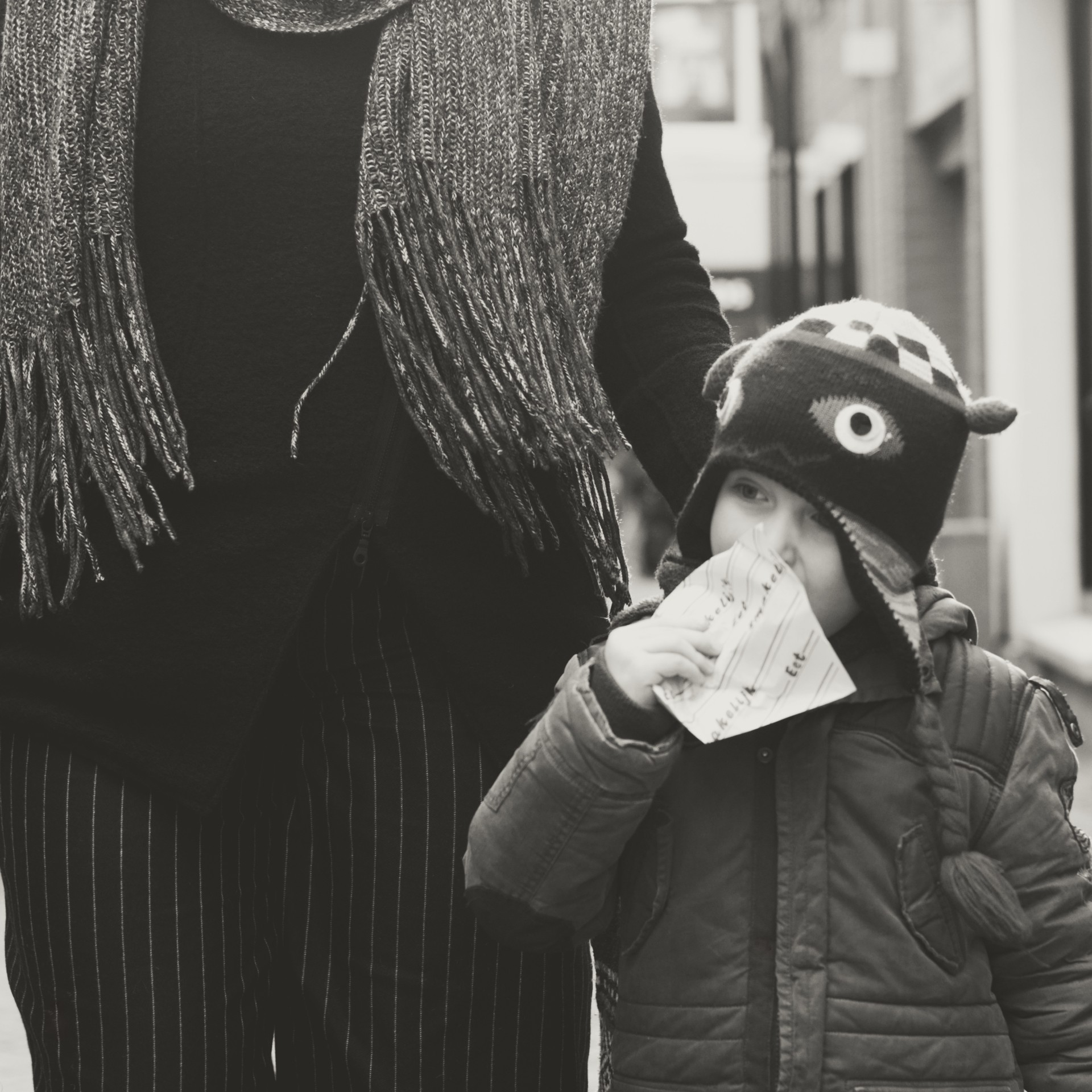 Candid black-and-white street photograph of an adult and child walking together, highlighting textured clothing, the child’s playful knit hat, and a small paper held to the child’s face, evoking a quiet moment of everyday life.