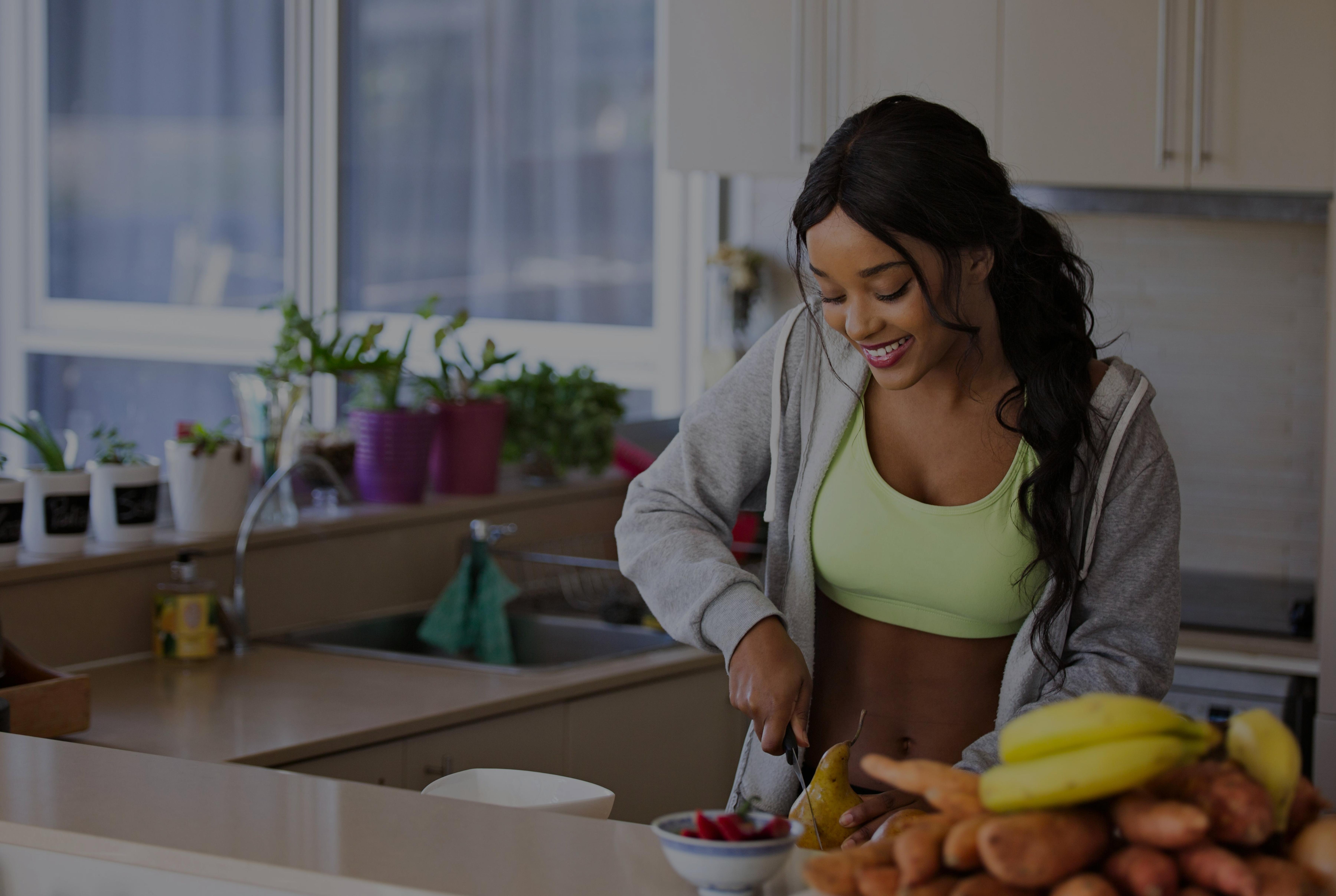 Smiling woman in sportswear cutting fruit in a bright kitchen surrounded by fresh produce and houseplants.