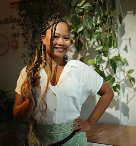 Uma empreendedora negra, com cabelos trançados, segura uma tesoura em uma das mãos.