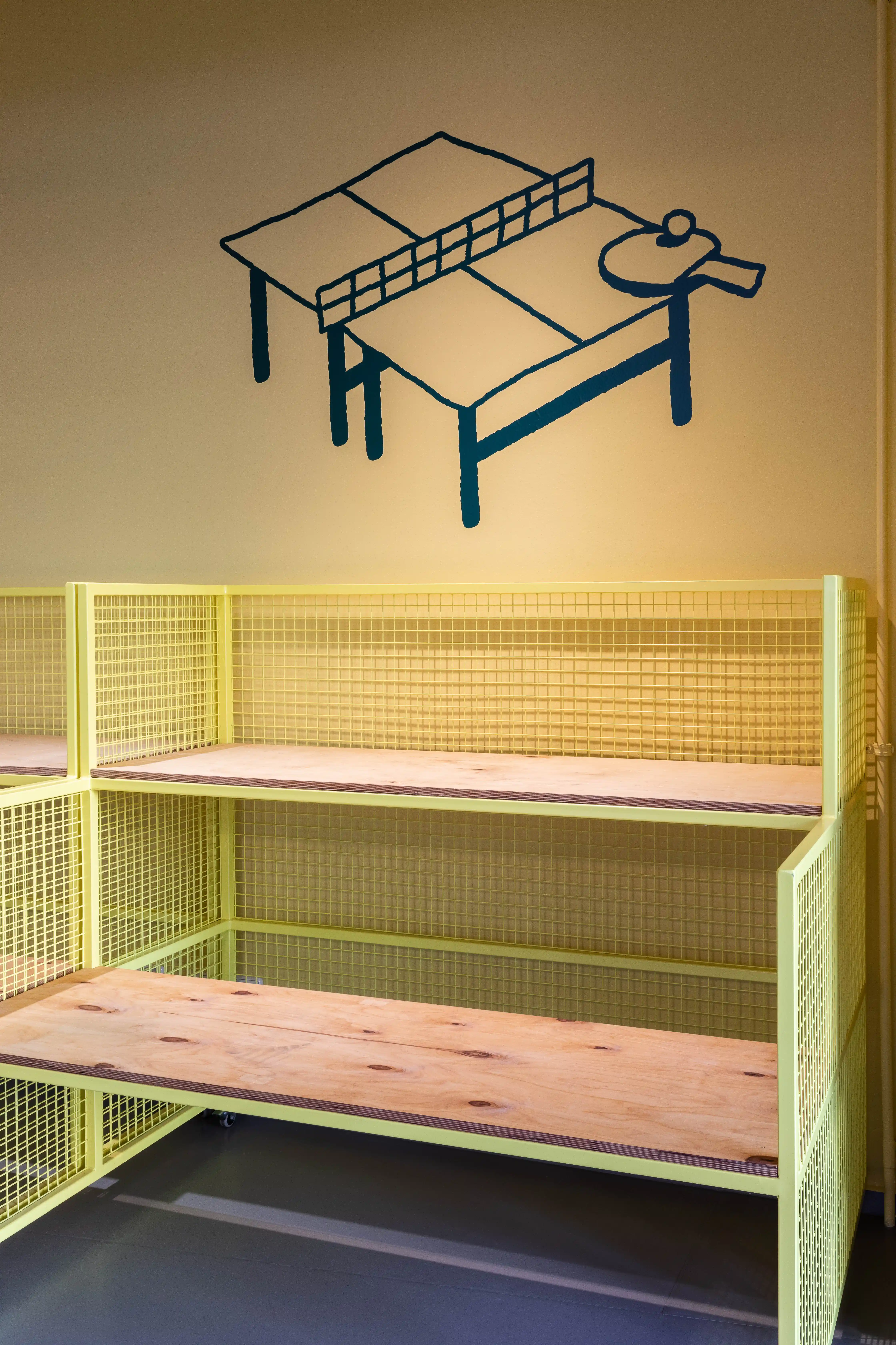 Yellow wire-mesh benches with wooden seats at DING DONG PING PONG beneath a dark blue mural of a table tennis table with paddle.