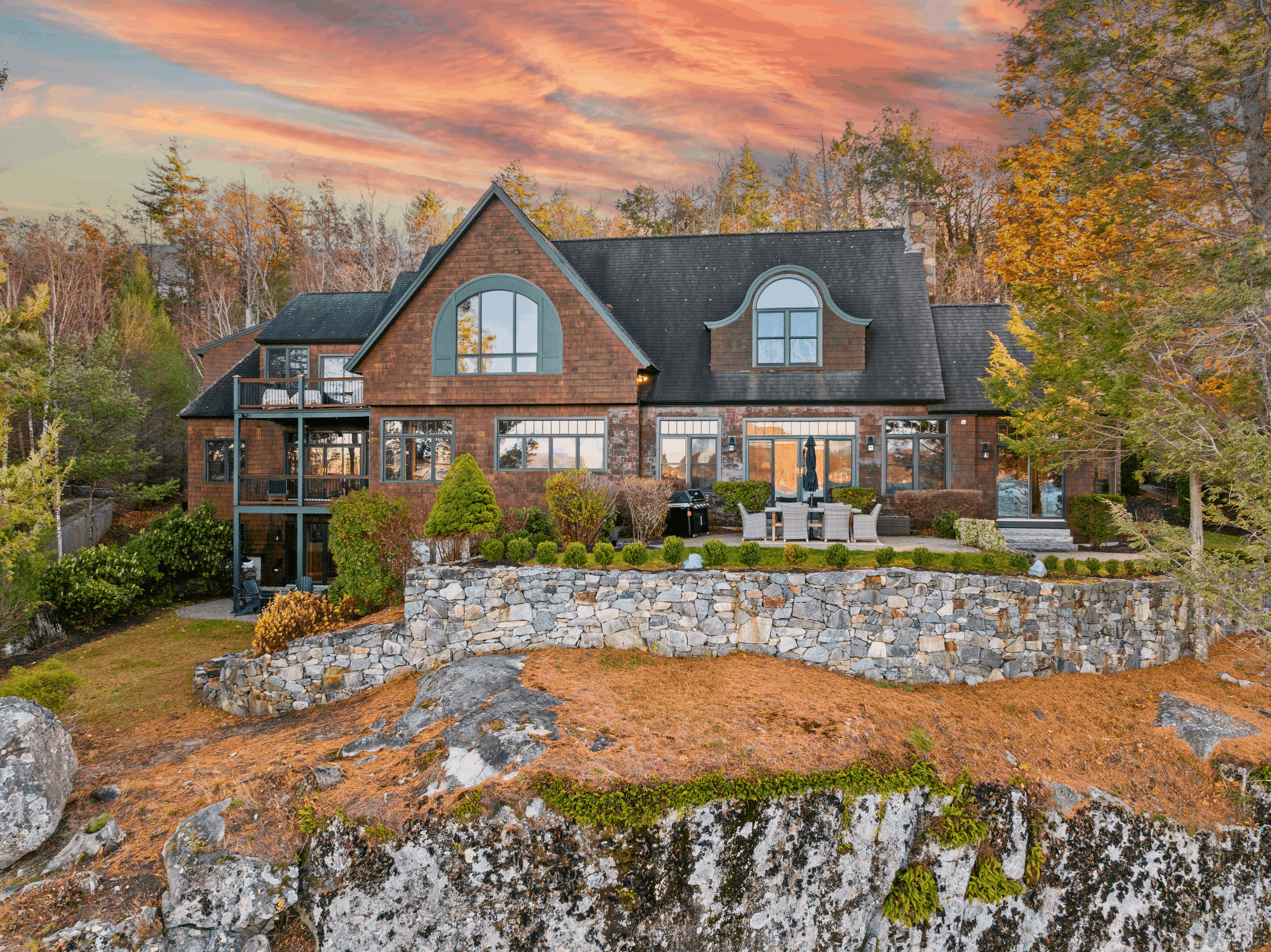 Luxury lakefront vacation rental home in Alton, NH on Lake Winnipesaukee with stone terrace, arched windows, and fall foliage at sunset — managed by Lodgism
