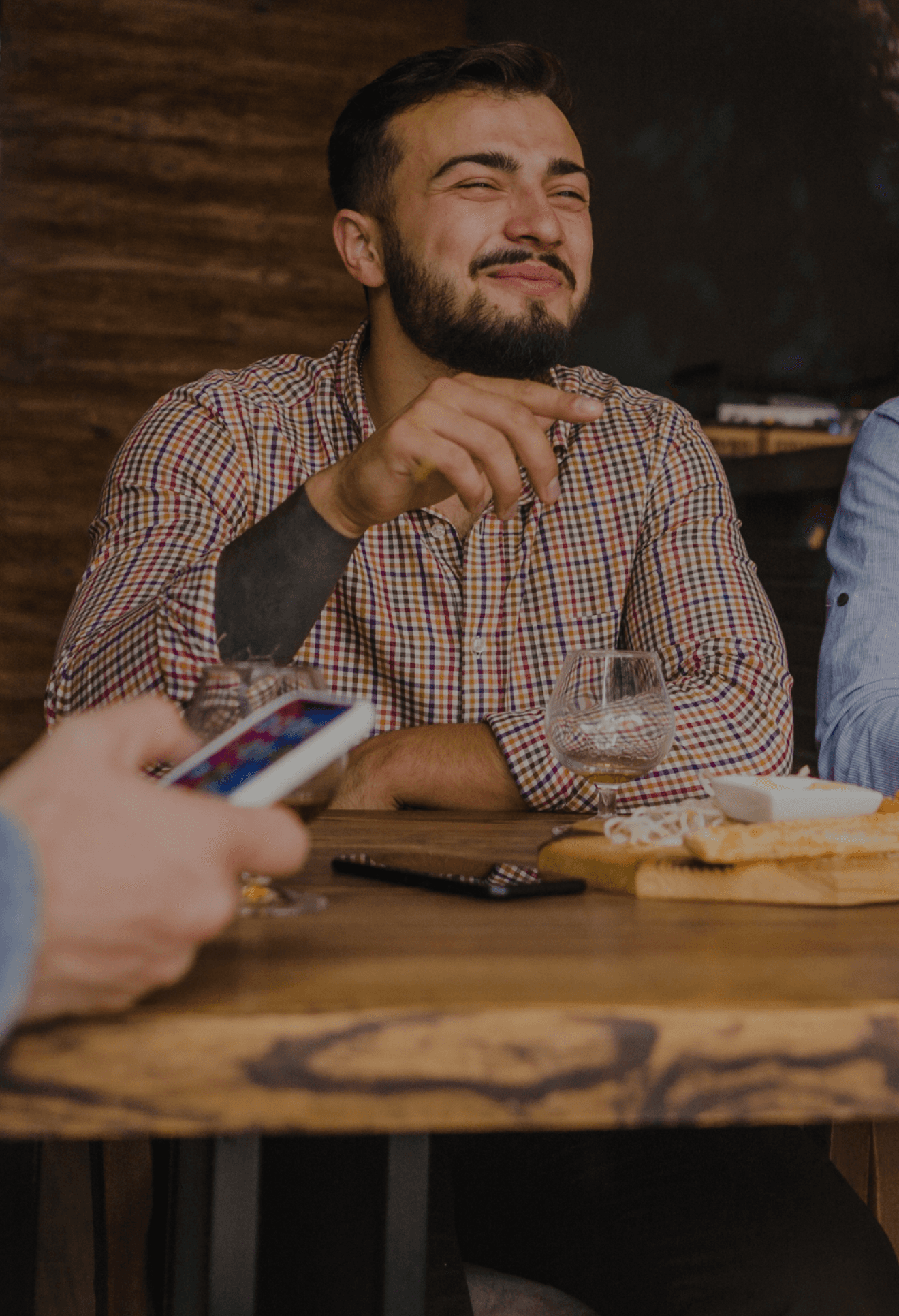 Un hombre está sentado en una mesa, sonriendo y sosteniendo una bebida en una mano. Parece estar disfrutando de un momento social, quizás compartiendo una comida o bebidas con amigos.