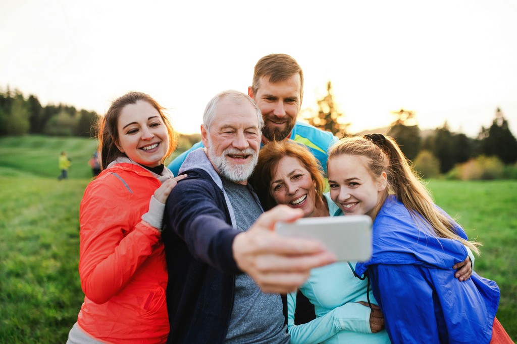 A cheerful multi-generational family takes a selfie together outdoors in a lush green field during sunset. The group, dressed in colorful jackets, includes grandparents, parents, and a young adult, all smiling and huddled closely. The scene captures the joy of family gatherings, outdoor activities, and creating lasting memories.