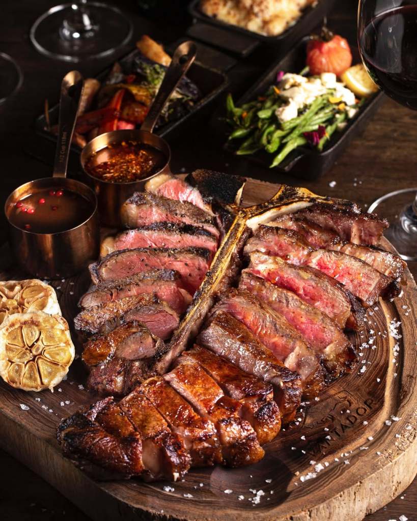 A beautifully presented assortment of gourmet steak cuts on a wooden platter, accompanied by roasted garlic, dipping sauces, and side dishes including vegetables and a salad, with a glass of red wine in the background.