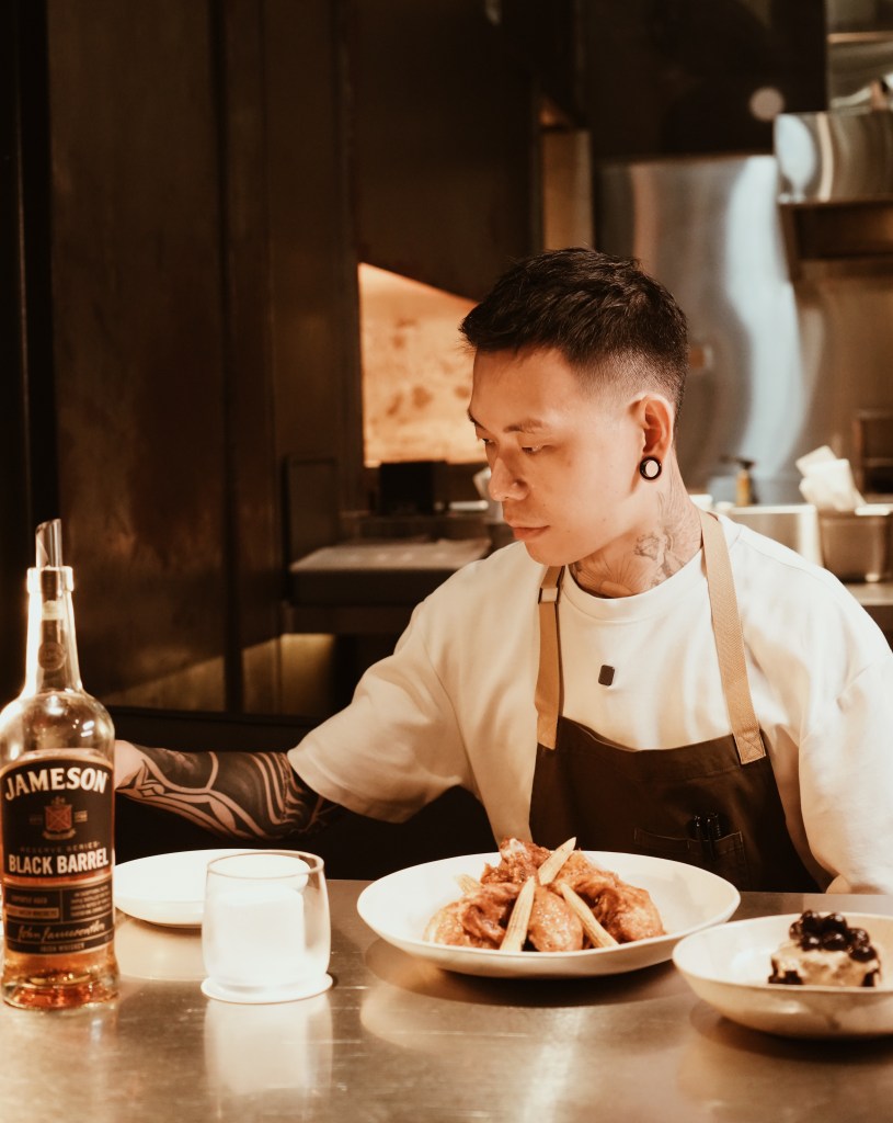 A chef in a restaurant setting, focused on plating a dish, with a bottle of Jameson Black Barrel whiskey on the table next to a plate of fried chicken and a dessert.