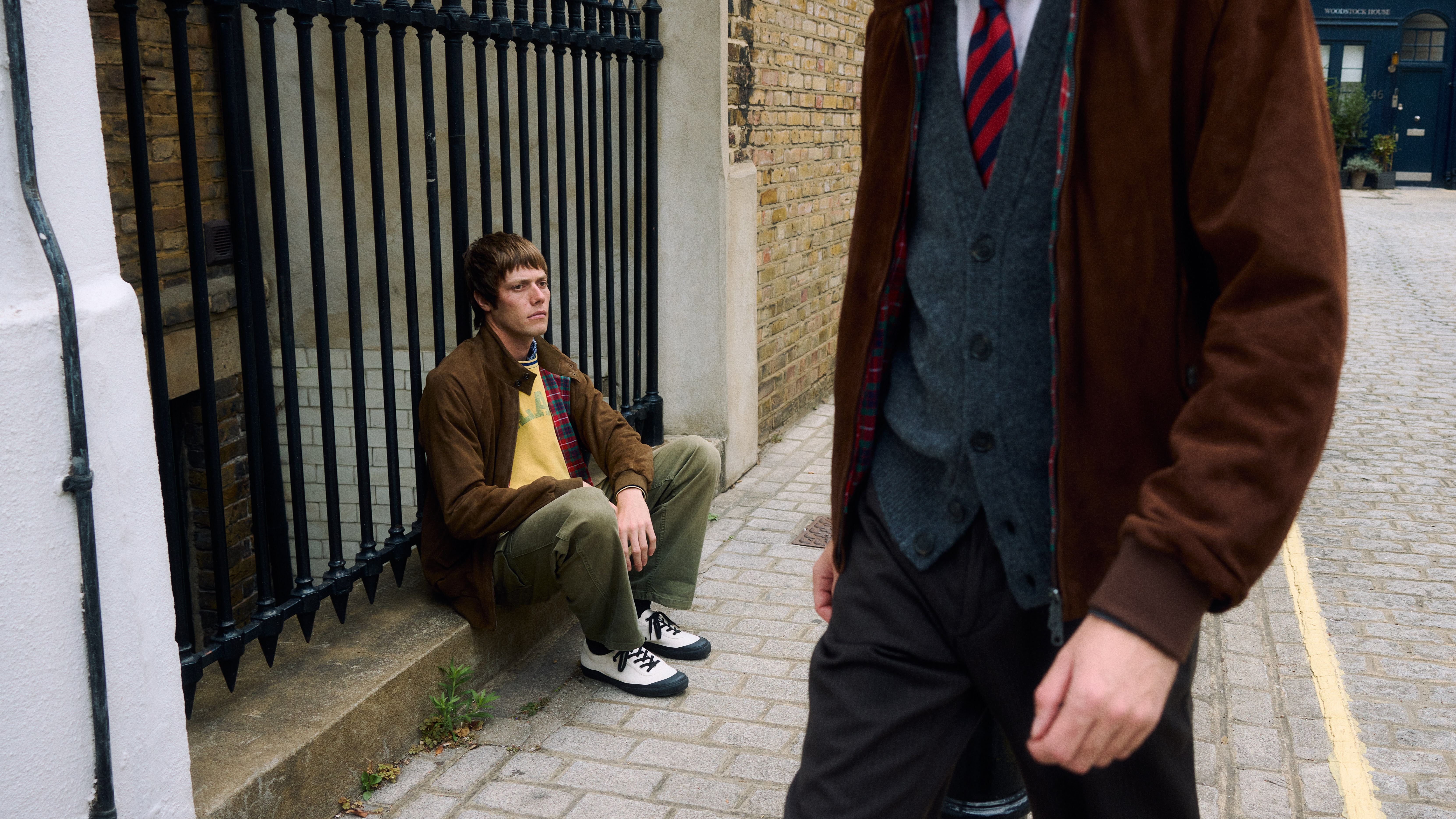 Au premier plan, le torse d'un homme vêtu d'un blouson en velours marron, sa camarade assis sur le trottoir porte ce blouson, un polo jaune et des baskets bicolores