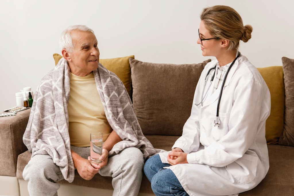 A friendly doctor in a white coat sits with an elderly man wrapped in a blanket, holding a glass of water, as they engage in a comforting and attentive conversation in a cozy living room setting.