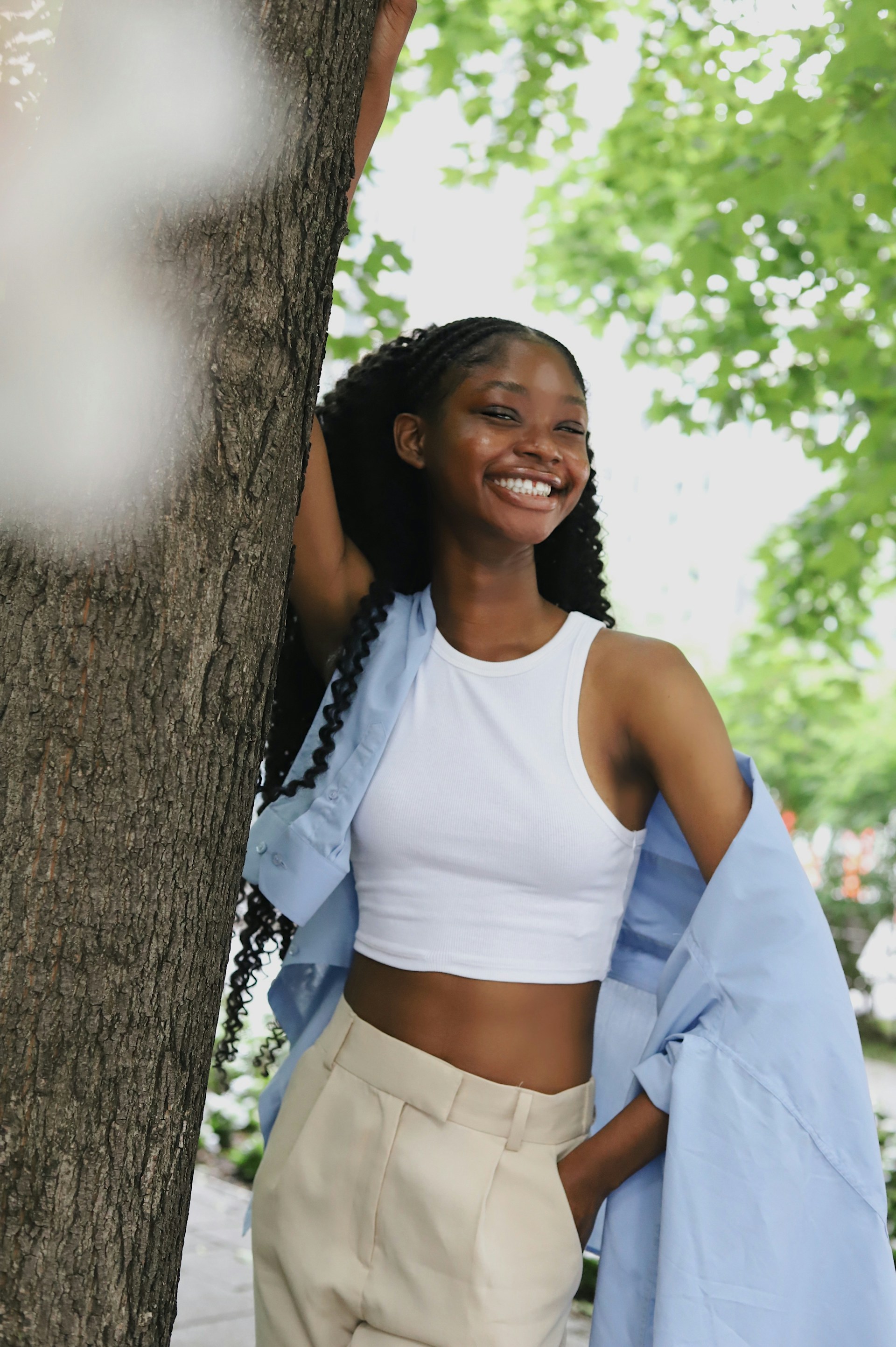 A woman with dark braids and long wavy hair leans against a tree trunk, smiling broadly. She is dressed in a white tank top, cream trousers, and a blue button-down shirt worn open.