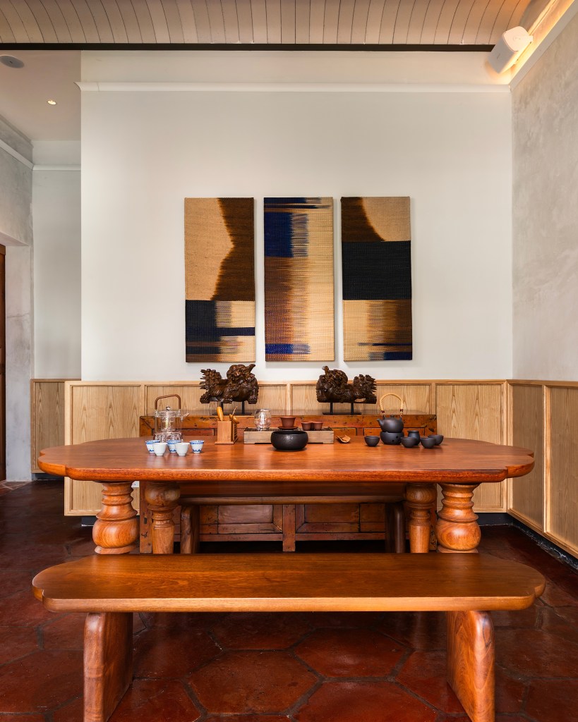 A cozy interior featuring a large wooden dining table with benches, accompanied by a tea set and decorative items. The wall showcases three abstract textile artworks, blending warm tones with blues.