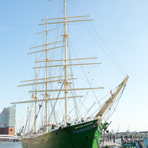 A large green and white sailing ship docked at a harbor with people walking nearby and modern buildings in the background.