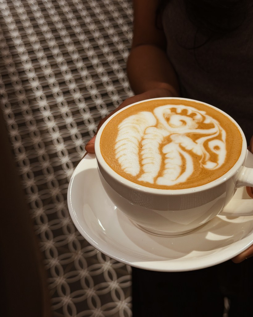 A person holding a white cup of latte with intricate coffee art on top, featuring swirls and leaf patterns, sitting on a white saucer against a patterned background.