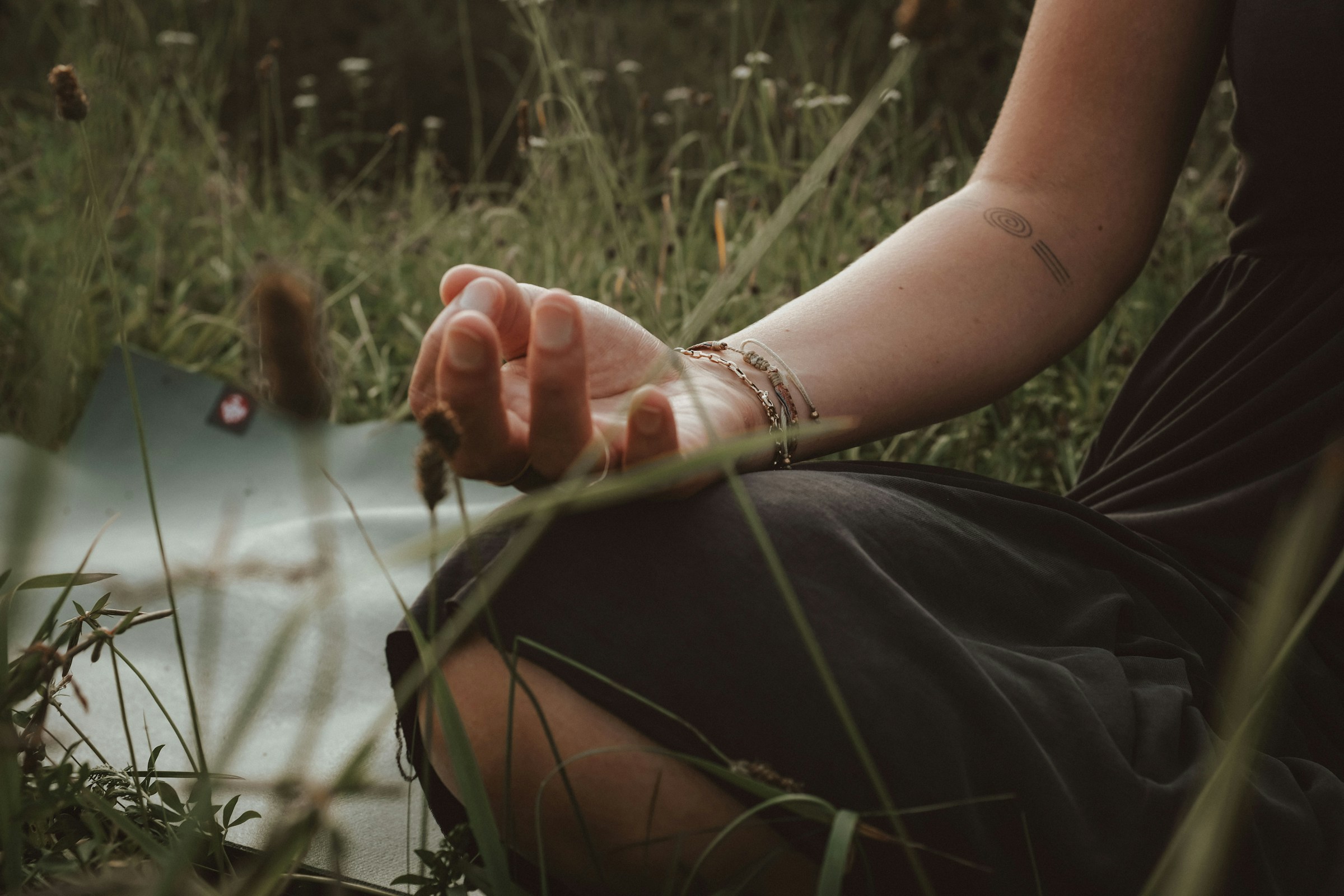 Woman in black dress sitting in grass with hand in mudra position, adorned with silver bracelets and a spiral tattoo on her arm, surrounded by tall blades of grass and flowers.