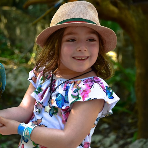 Uma jovem vestindo um vestido floral e um chapéu, sorrindo ao ar livre. Ela tem uma pulseira azul no pulso esquerdo.