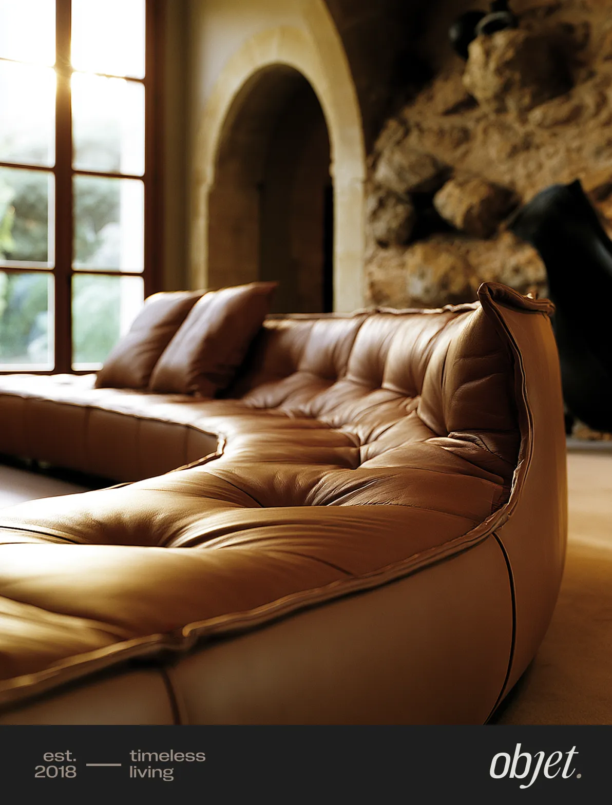 Editorial photograph of a cognac leather tufted sofa in a warm Mediterranean-style room with stone arched walls and sunlight filtering through a tall window, with the "objet." wordmark and "est. 2018 — timeless living" label along the bottom edge