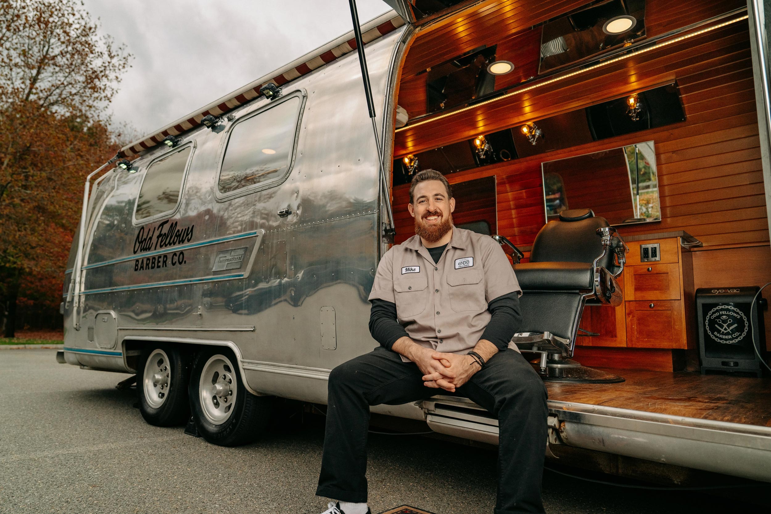 A bearded man in a tan work shirt is sitting on the step of a shiny, vintage-style aluminum travel trailer that has been converted into a mobile barbershop. The side of the trailer is labeled "Odd Fellows Barber Co." The interior, visible through the open side, shows a barber chair and wood paneling. The man is smiling and looking at the camera.