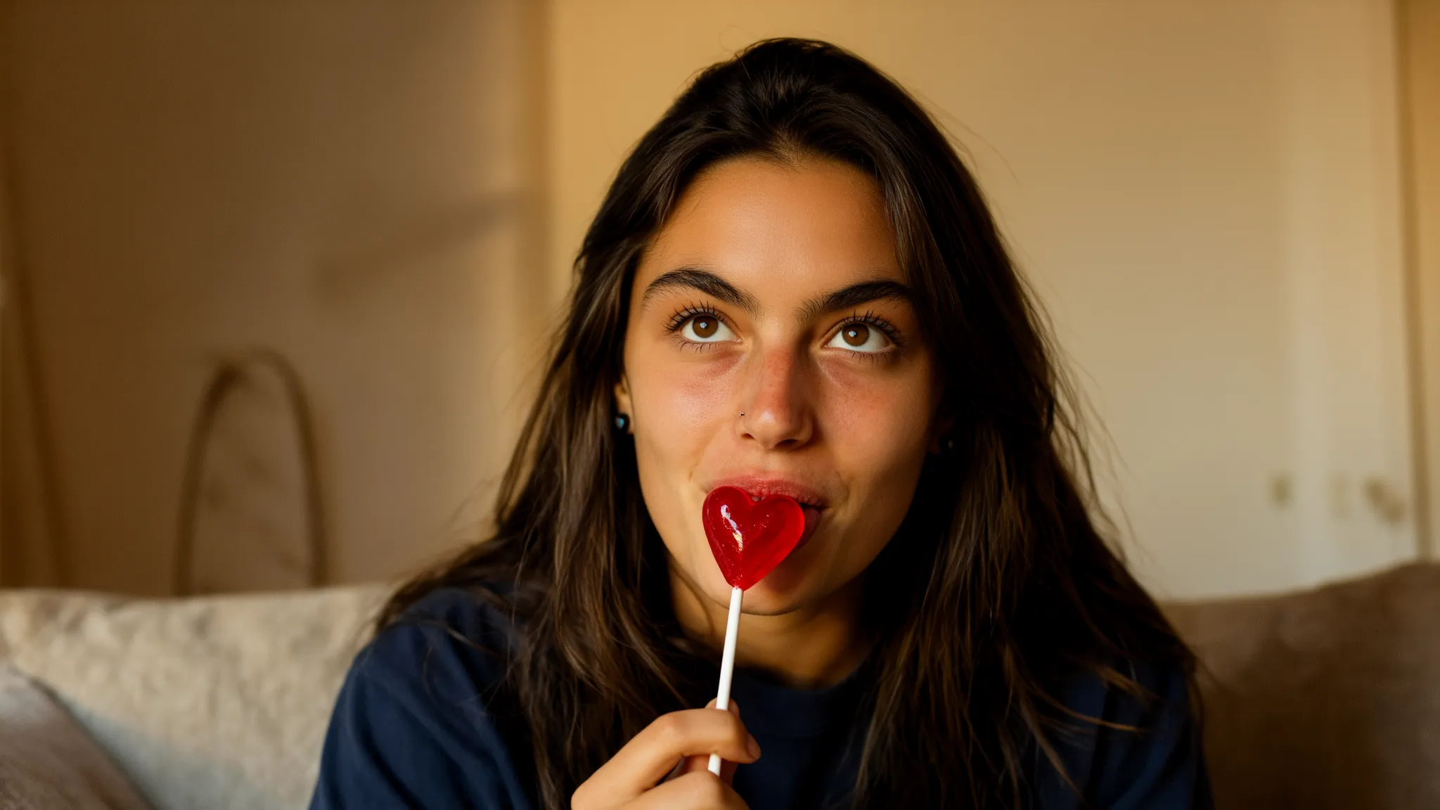 Weibliches KI-Model im warmen Zimmerlicht mit langem dunklem Haar, das einen roten Herz-Lolli genießt und verspielt in die Kamera blickt.