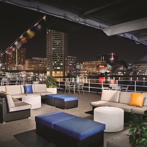Rooftop lounge area with modern seating, overlooking a city skyline and water at night, decorated with string lights.