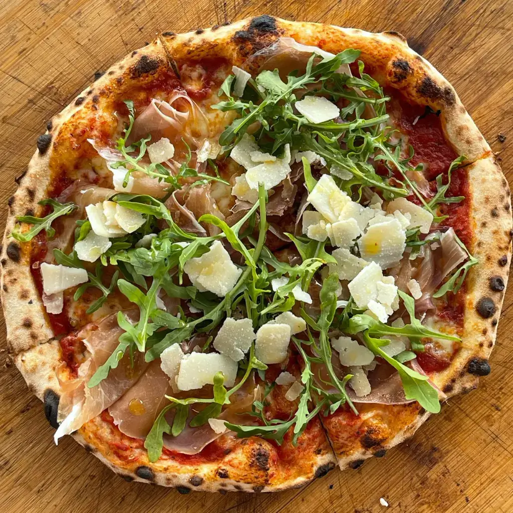Aufgeschnittene Pizza mit Parmaschinken, Rucola und Parmesan auf Holzbrett Pizza Dielsdorf