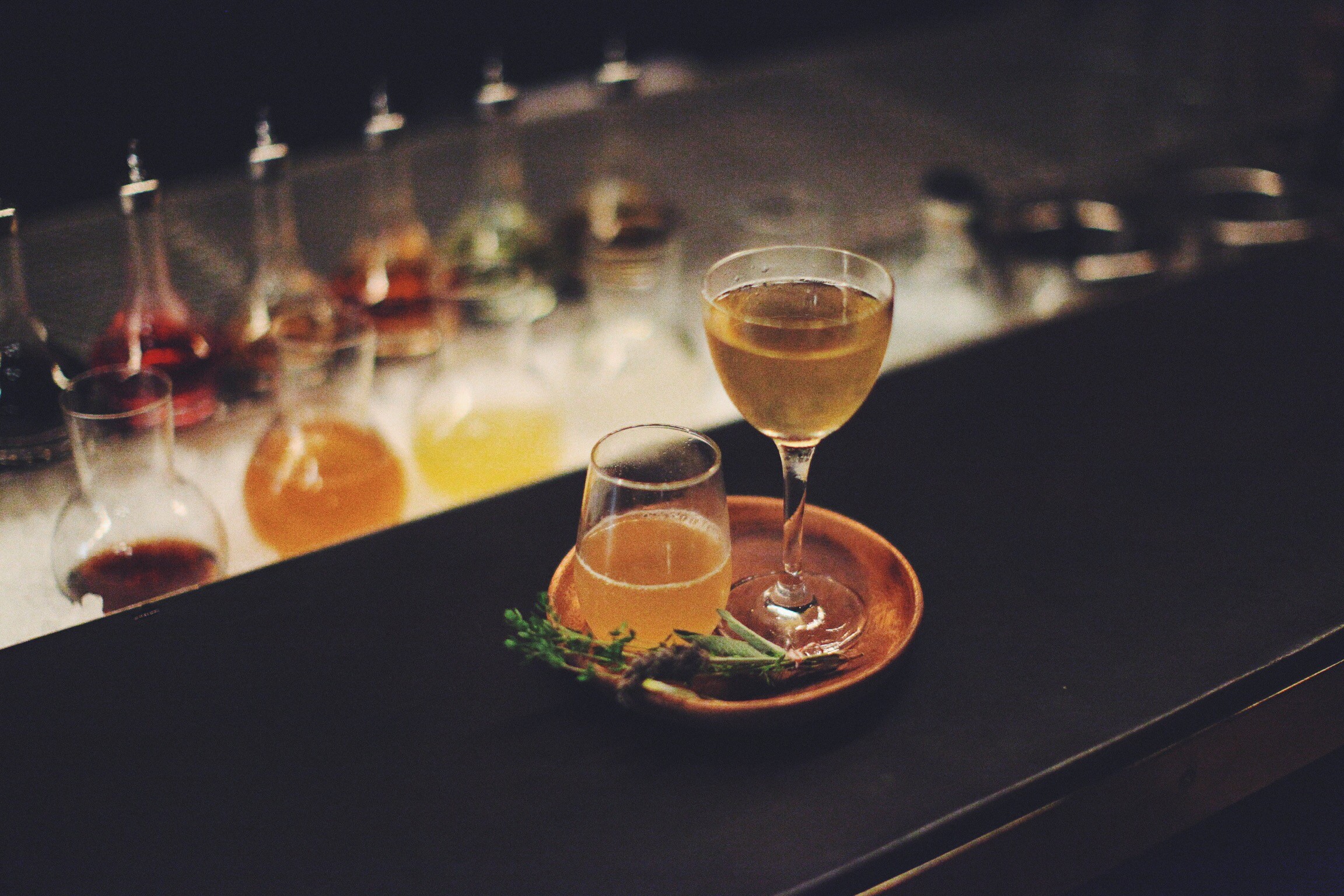 Two amber cocktails on a round tray at a dark bar, with bottles and glassware softly blurred behind.