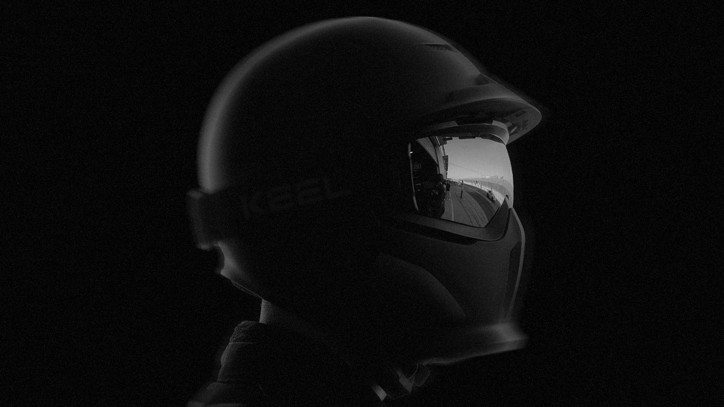 Black and white side profile of a helmeted driver, with reflective visor and minimal lighting against a dark background.