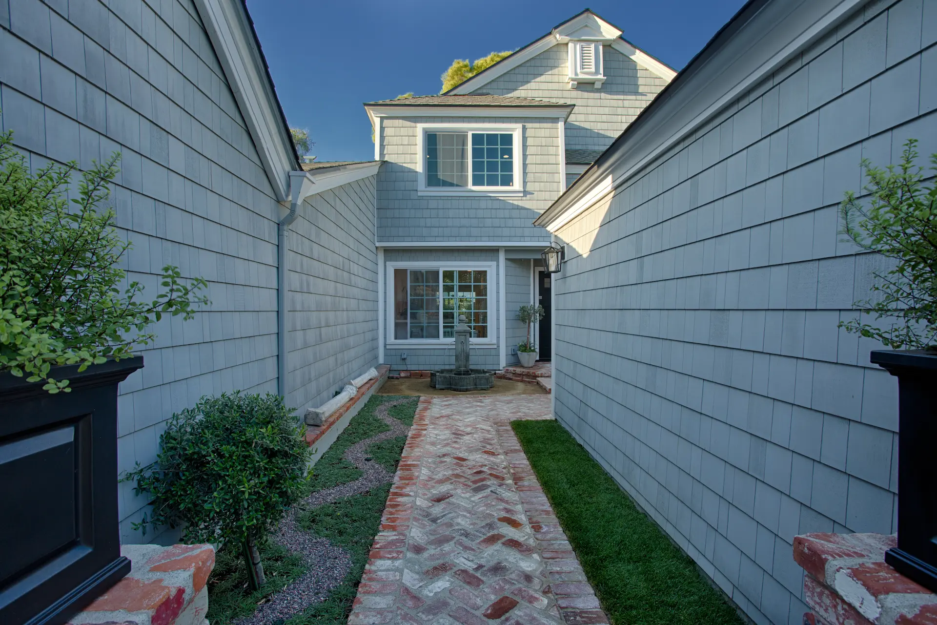 Narrow path to a Cape Cod home's back unit, flanked by grey walls and green grass, brick accents on the path. From Dana Point Cape Cod remodel by Chris Darnall.