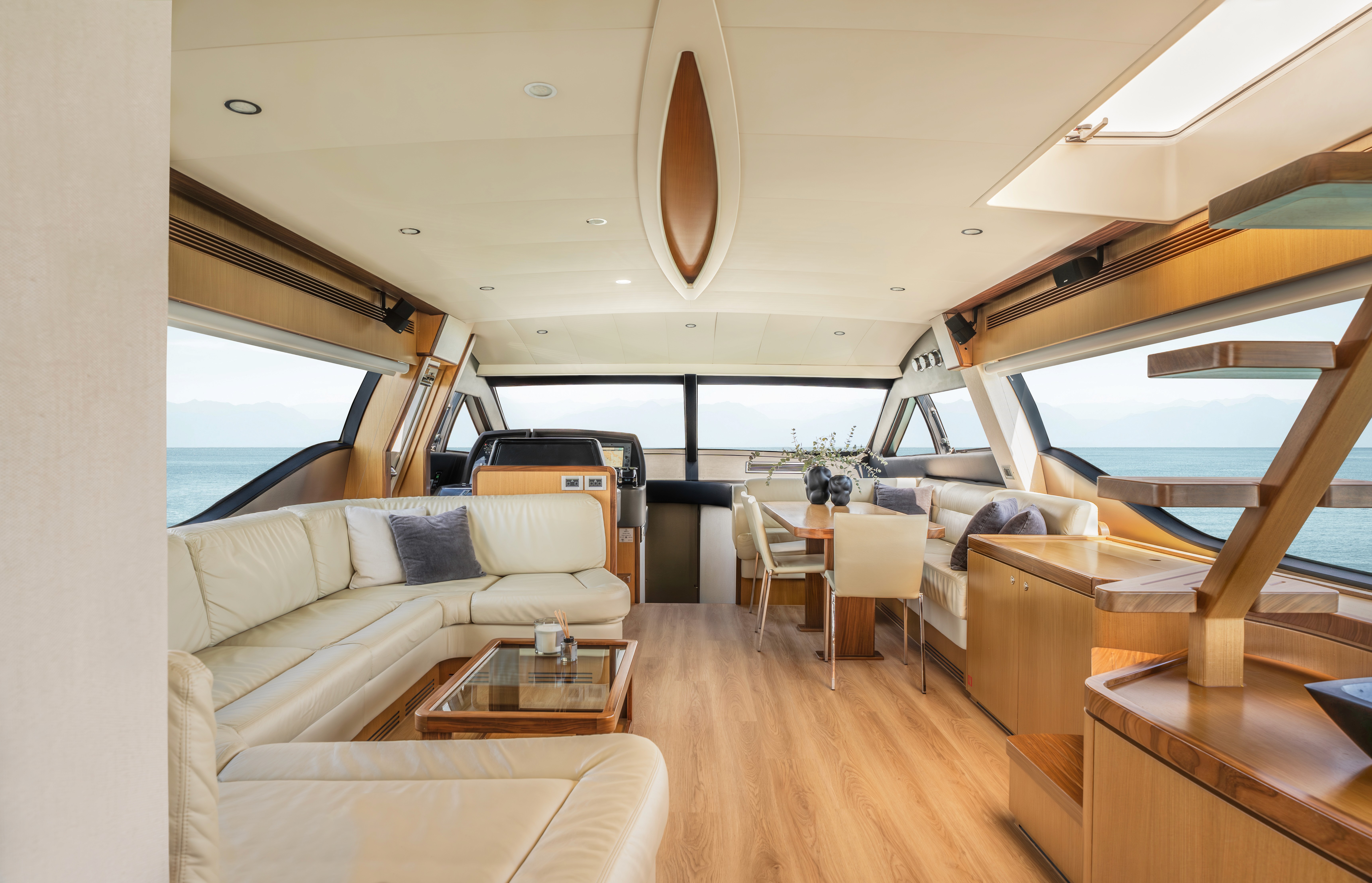 Luxurious yacht interior cabin with warm wood paneling, cream leather seating, and large windows overlooking calm blue waters.
