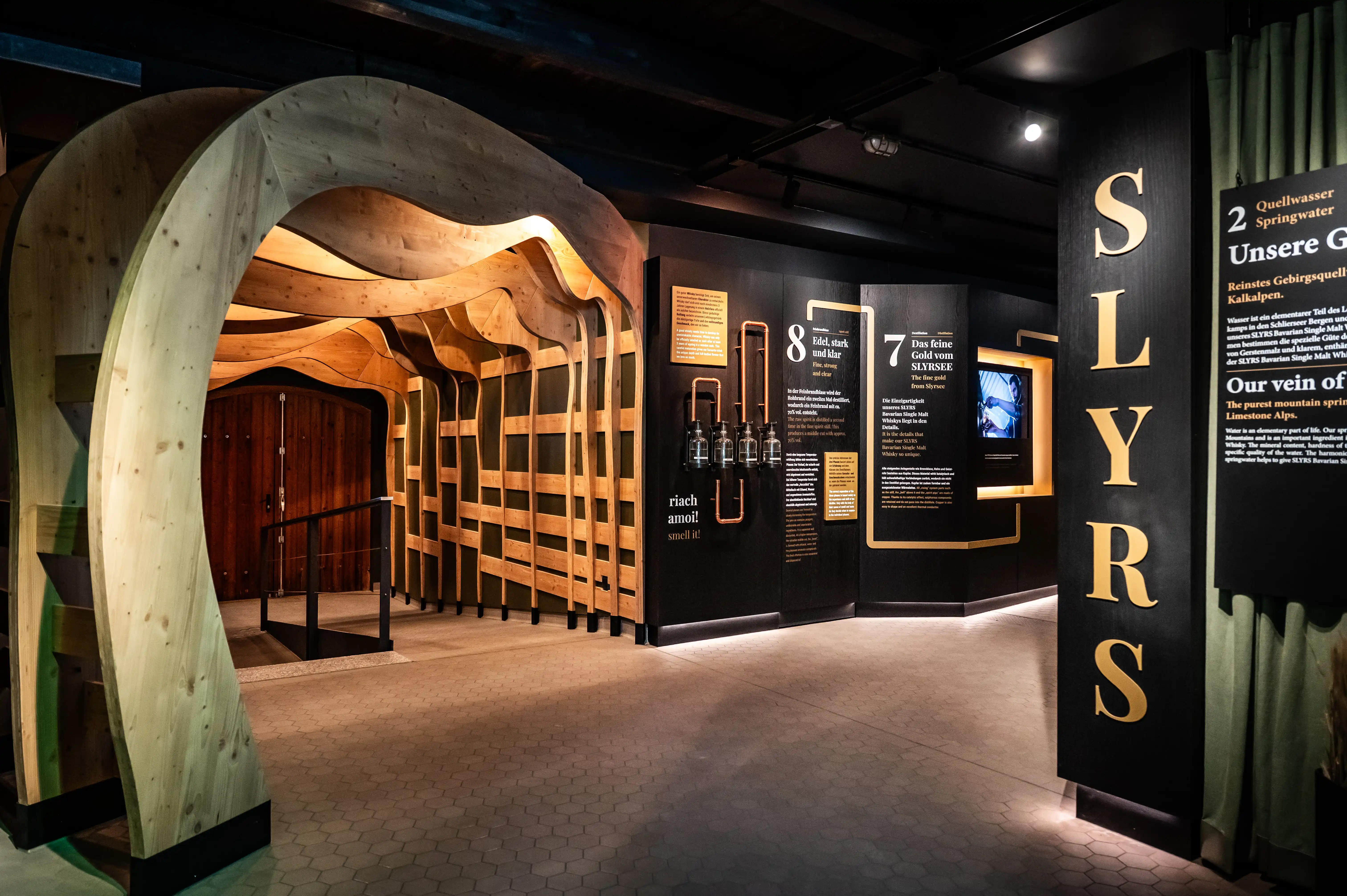 Exhibition room of the SLYRS Experience Centre with curved wooden arch, golden vertical logo and dark info walls.