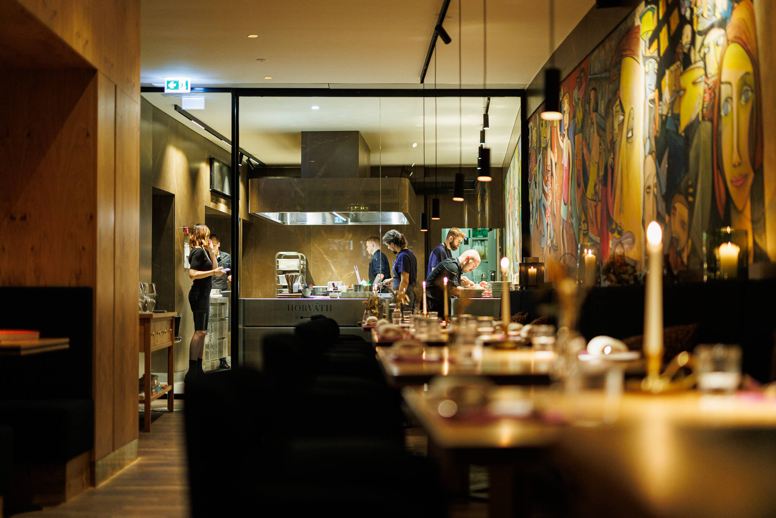 Blick durch den Gastraum auf die offene Küche; gedeckte Tische mit Kerzen im Vordergrund.