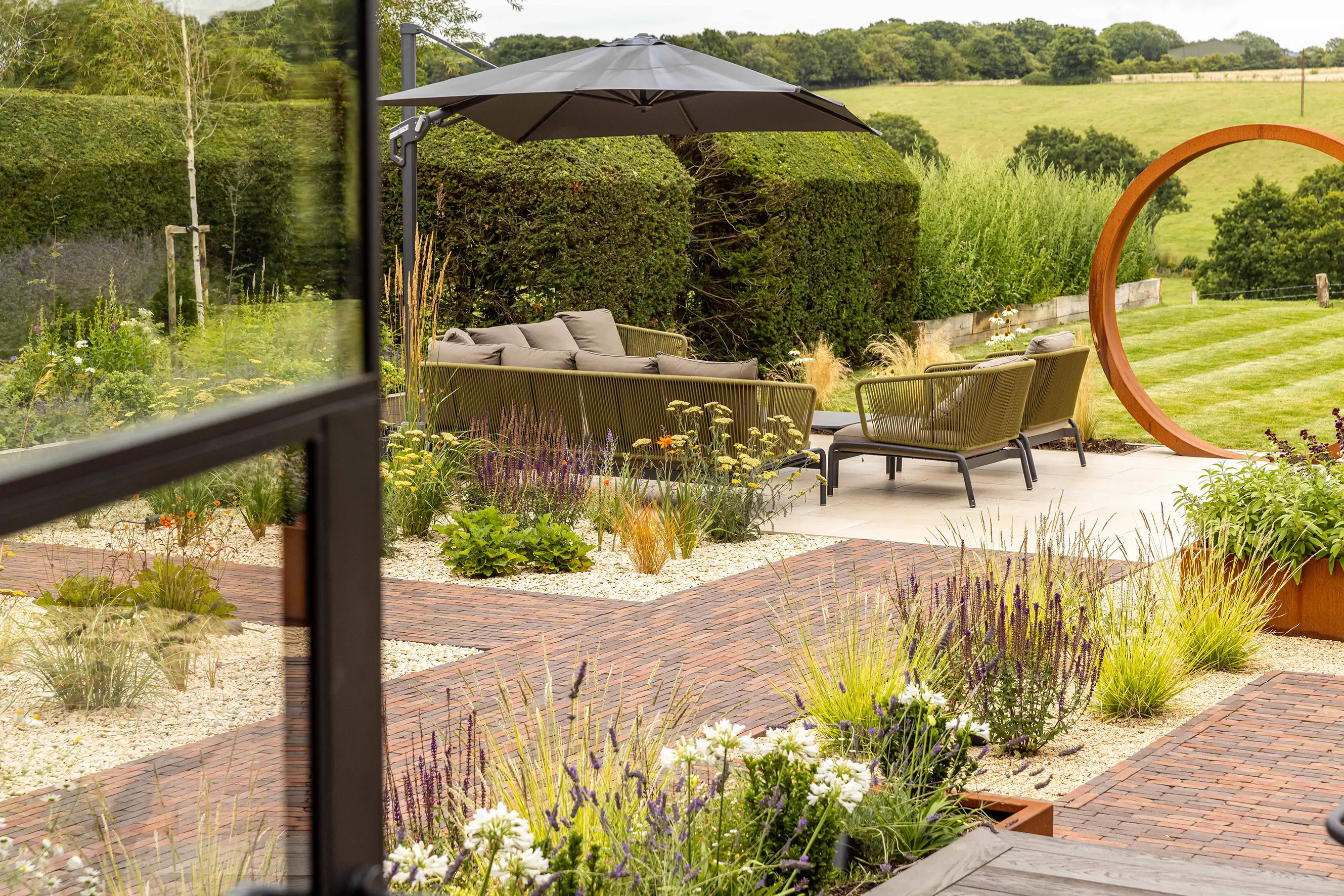 A landscaped garden view with pathways, seating areas, trees, and open fields in the background.