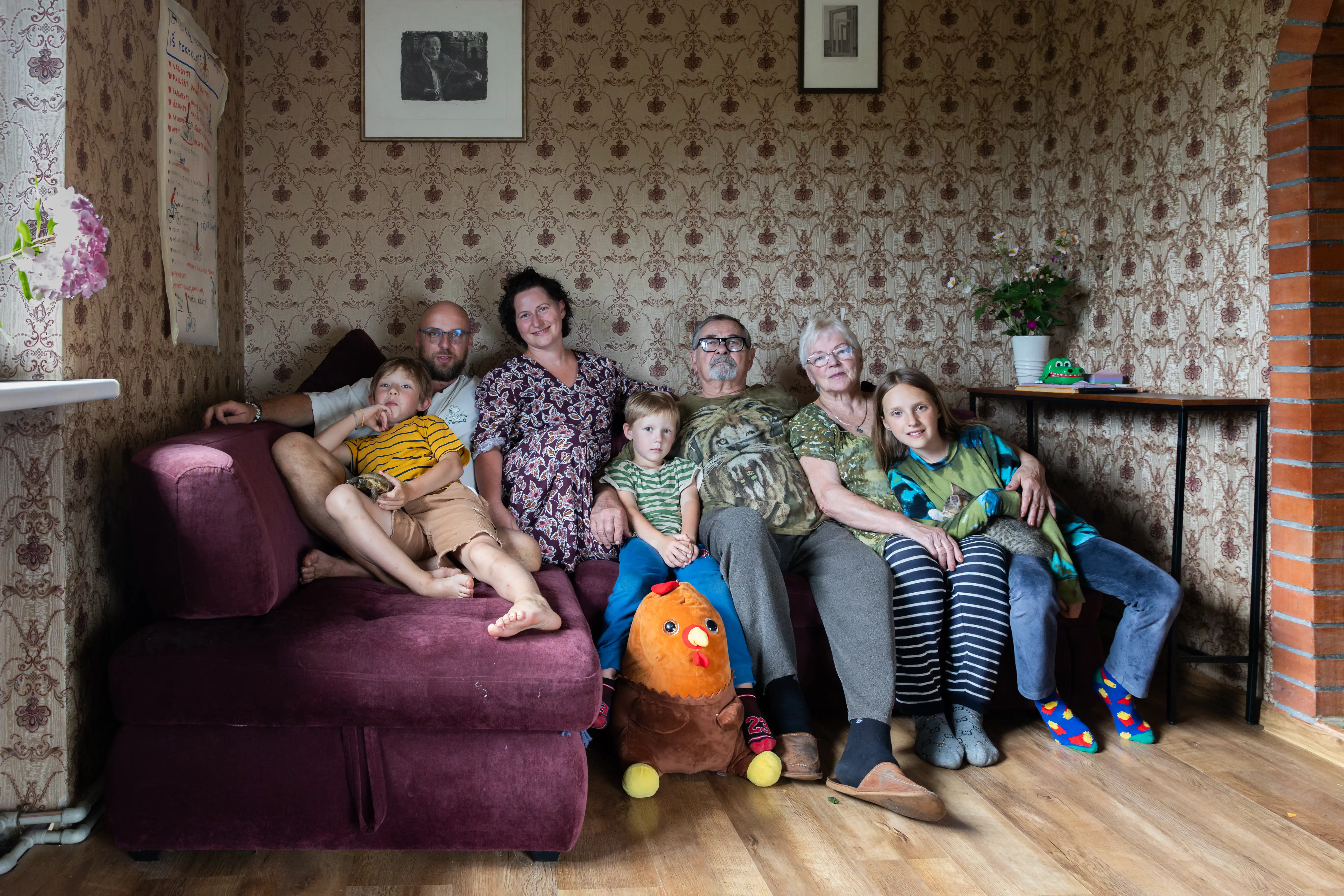 Rodzina wielopokoleniowa siedząca na kanapie w przytulnym pomieszczeniu. W skład grupy wchodzą dorośli i dzieci, a w tle widoczne są kwiaty na stole i zdobione ściany.