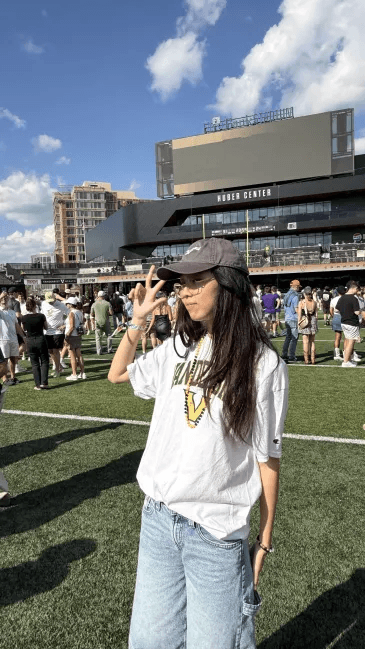 Una persona con una camiseta negra y pantalones de color claro está de pie en la cima rocosa de una montaña al atardecer, sosteniendo una cámara frente a su cara para tomar una foto. Un mar de nubes se extiende detrás de ellos, iluminado por la luz dorada del sol poniente.