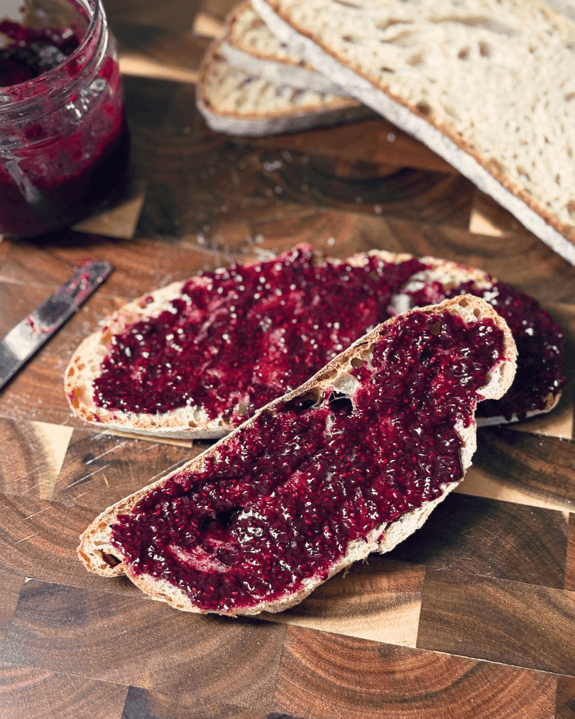 Blueberry chia jam spread on sliced sourdough bread, with a jar of jam and extra bread in the background on a wooden board.
