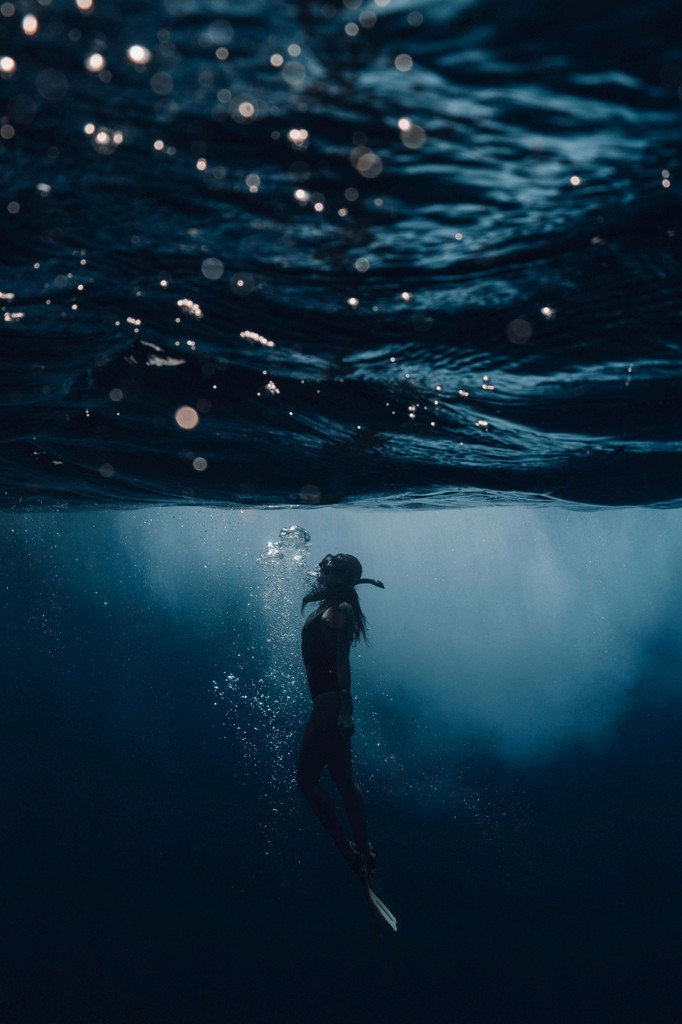 Close-up portrait of a person wearing a gray insulated hooded jacket with the hood up, showing their face framed by the hood against a light background.