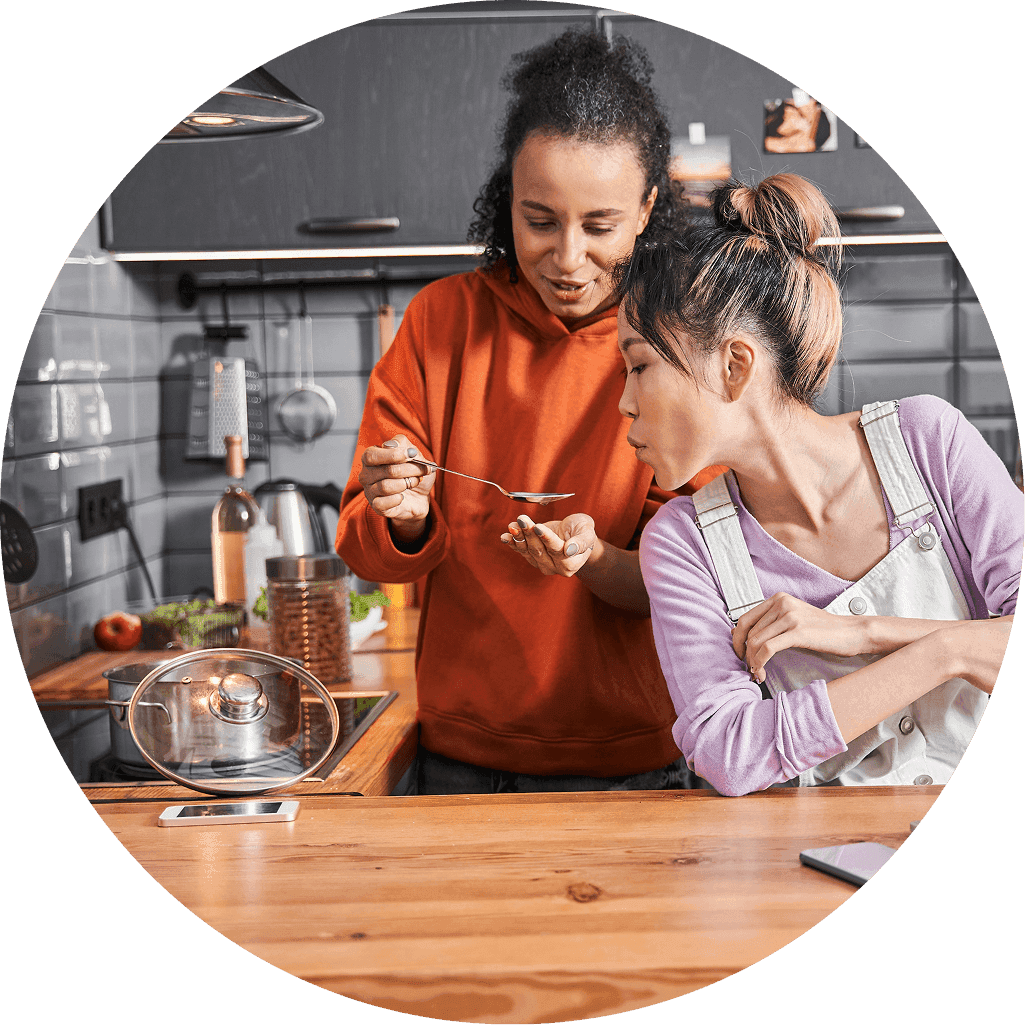 Two women cook together in a modern kitchen as one tastes food from a spoon while the other watches