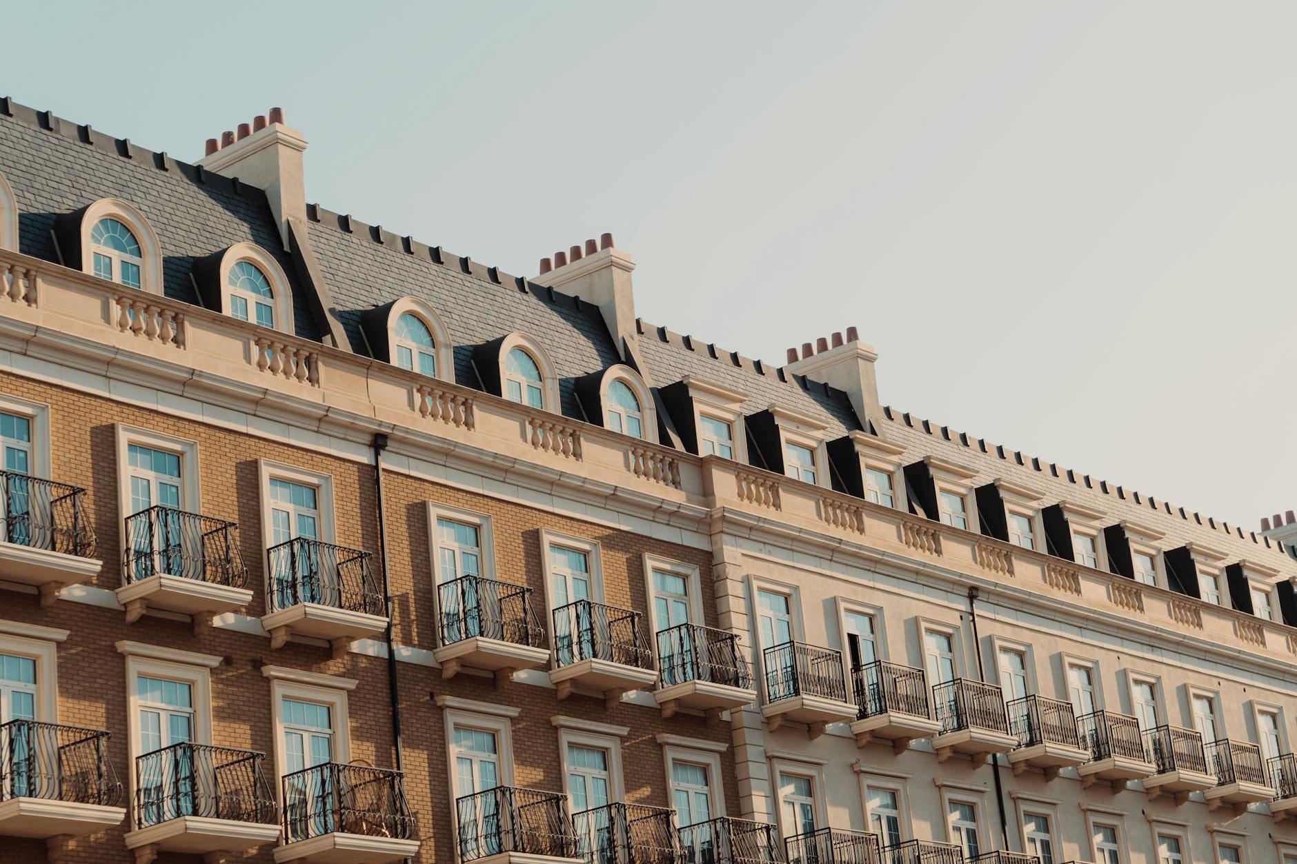 Upper floors of a Victorian apartment block with curved dormer windows, iron balconies and chimneys against a pale sky