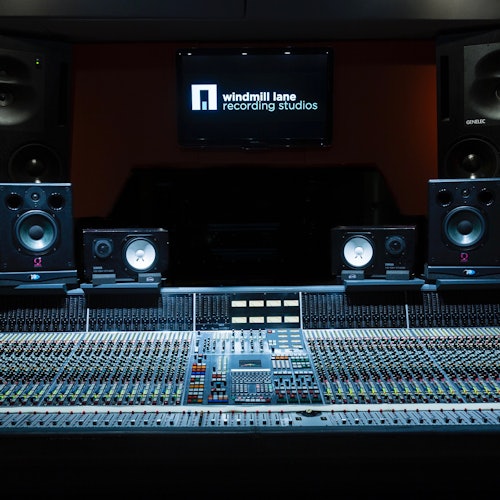 Close-up of a recording studio mixing console with multiple dials and switches, large speakers, and a screen displaying "Windmill Lane Recording Studios."