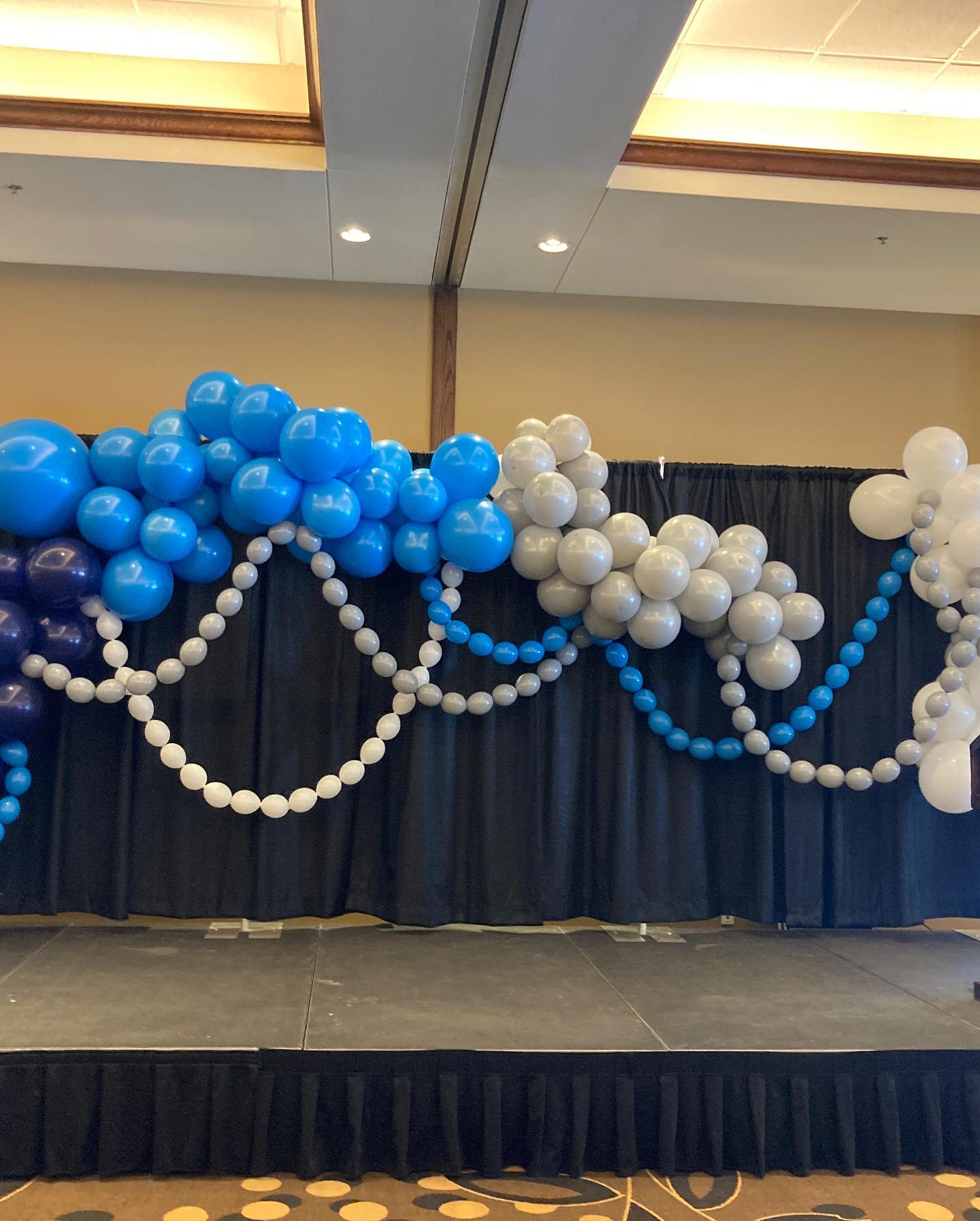 A colorful balloon arch featuring vibrant blue, white, and navy balloons decorates a stage area against a black curtain backdrop, perfect for a festive event or celebration.