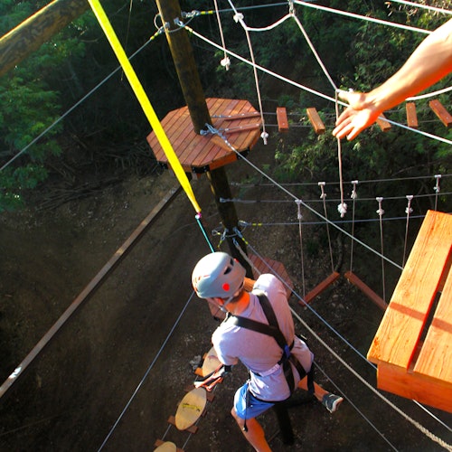 一个戴着头盔和安全带的人在距离地面较高的绳索课程中穿梭,周围环绕着树木和木质平台。