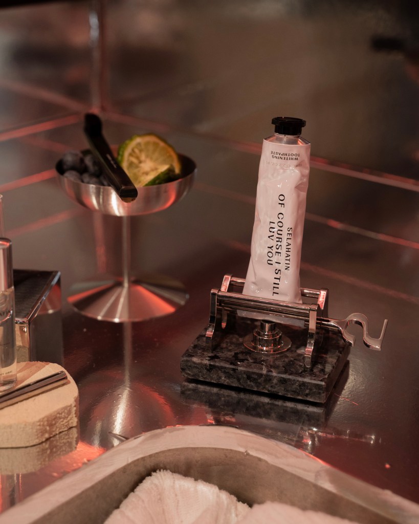 A stylish countertop setup featuring a tube of hand cream in a metal holder, a small bowl with a slice of lime and some blueberries, a glass bottle, and a towel.