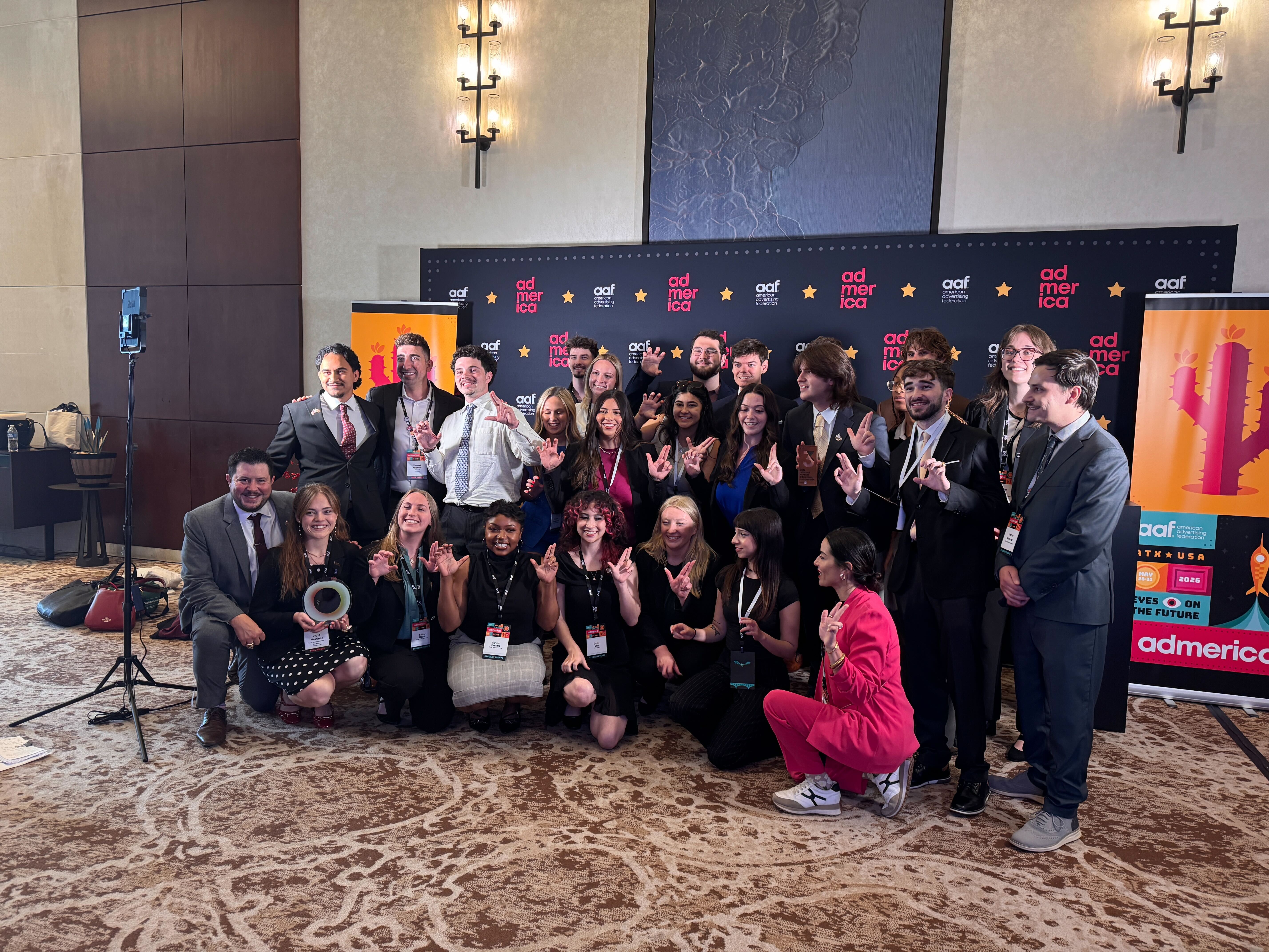 The winning student team from Texas State University celebrates at the AAF AdAmerica competition, posing for a group photo in business attire. Arranged in standing and kneeling rows, many are smiling, waving, or making playful hand gestures, and several hold trophies. Behind them is a navy step-and-repeat backdrop patterned with the 'AdAmerica' logo, the American Advertising Federation name, and gold stars. Tall, colorful event banners stand on either side, featuring cactus and rocket illustrations, 'ATX USA,' the dates 'May 28–31, 2026,' and the theme 'Eyes on the Future.' The setting is a carpeted ballroom with wall sconces.