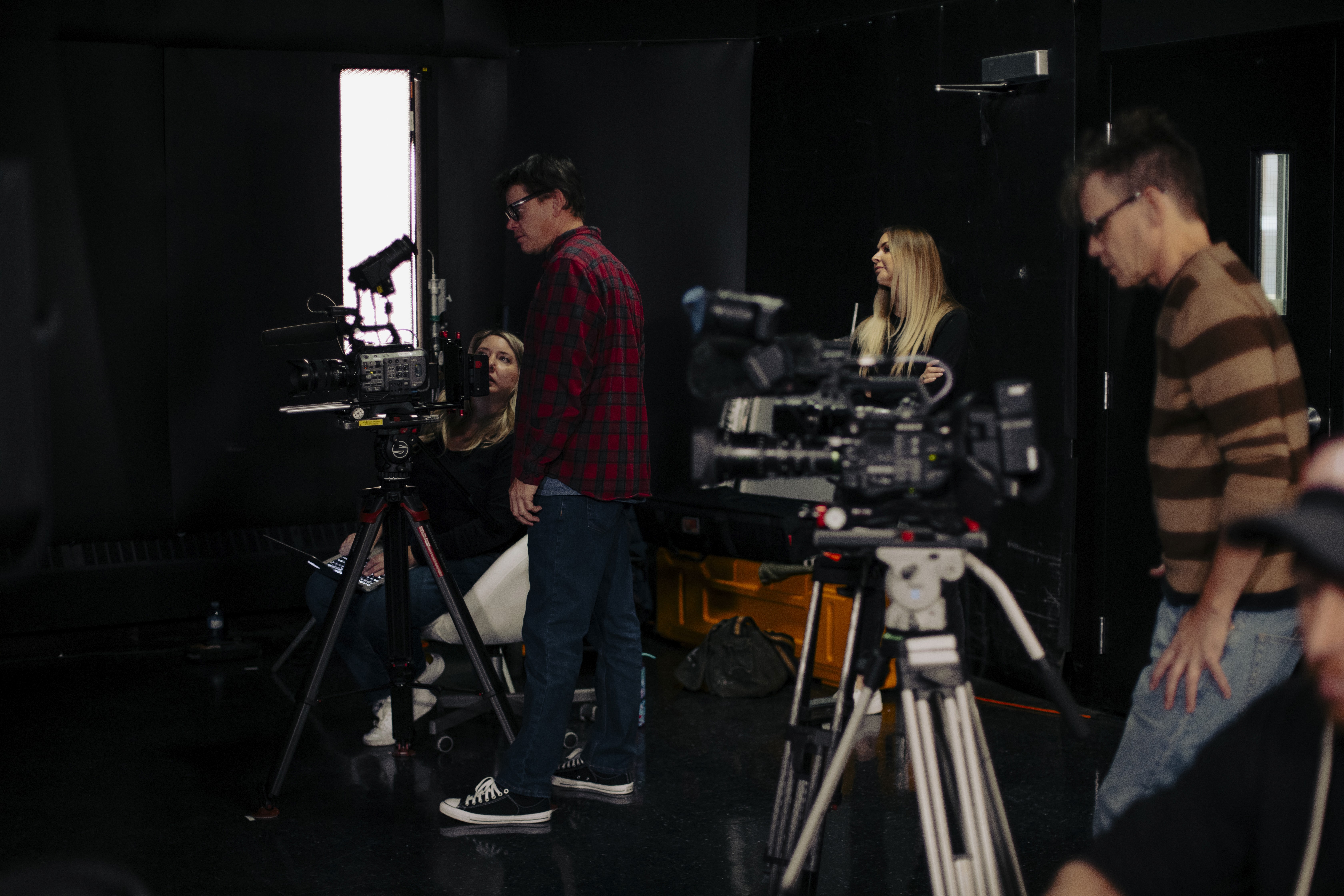 Two camera operators in a dimly lit studio adjust a large video camera. They wear headsets and caps, focusing intently. Mood is professional and focused.