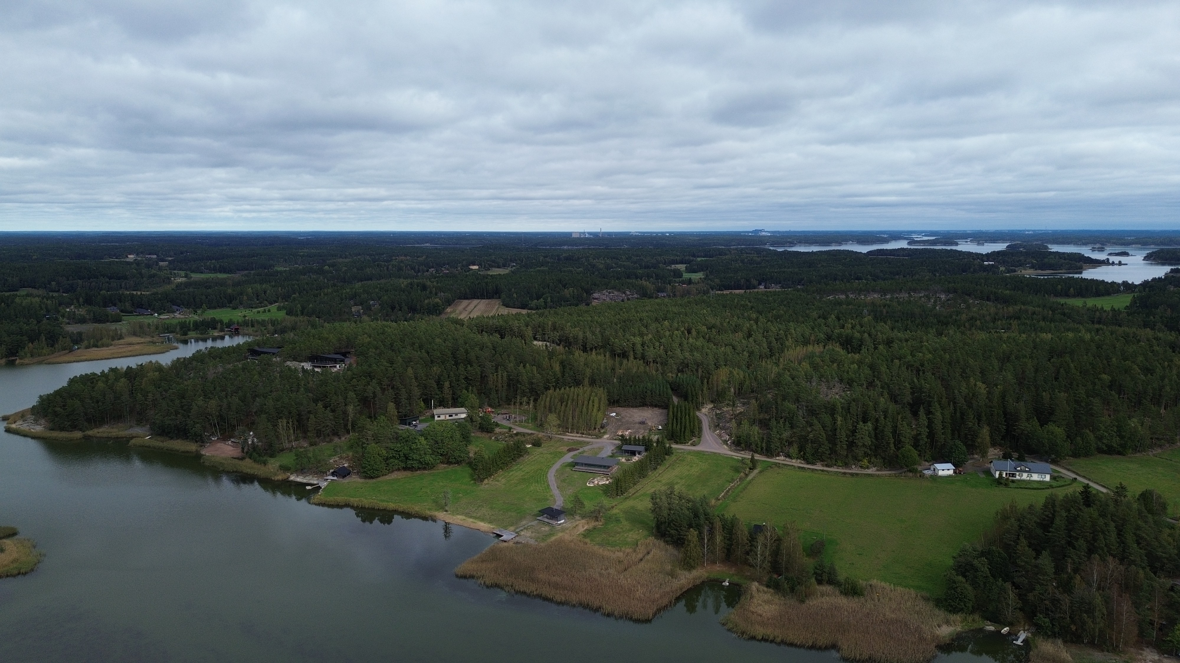 A plot depicted from a distance, featuring a house, a sauna, and a carport. The plot is surrounded by spruce trees.