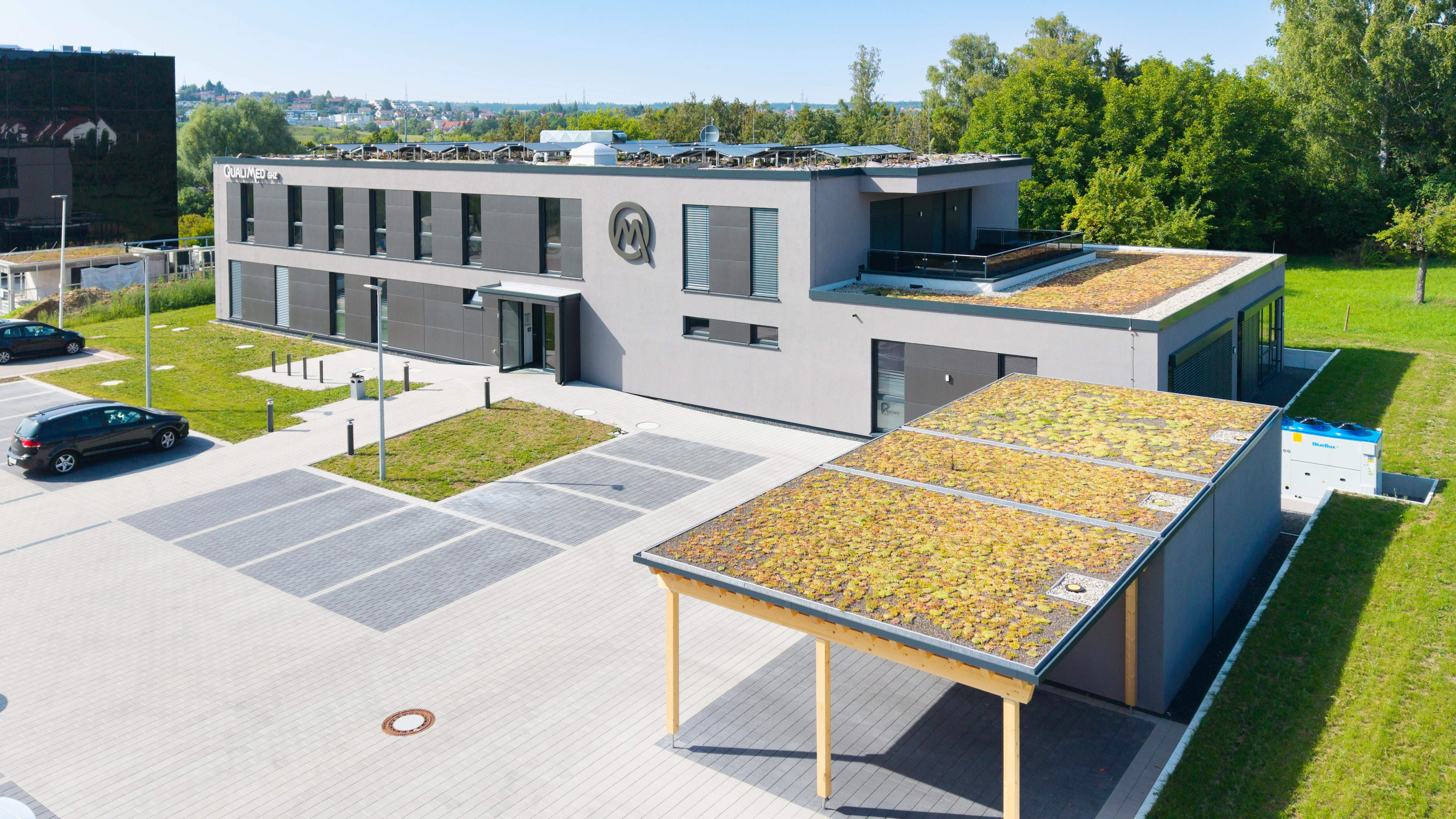 Modernes Bürogebäude mit Grünflächen, Photovoltaik auf dem Dach und einer Besucherparkplatzüberdachung mit Pflanzenbewuchs.
