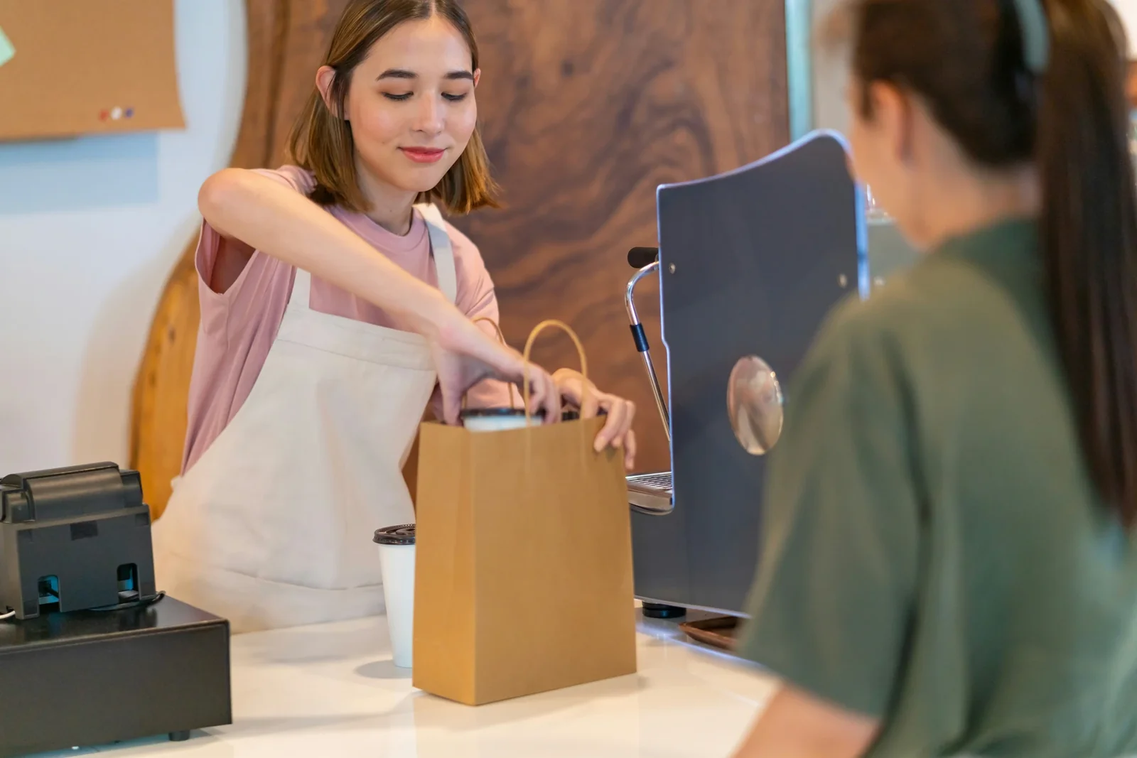 Retail employee hands customer a brown paper shopping bag at checkout counter with tablet and register visible.