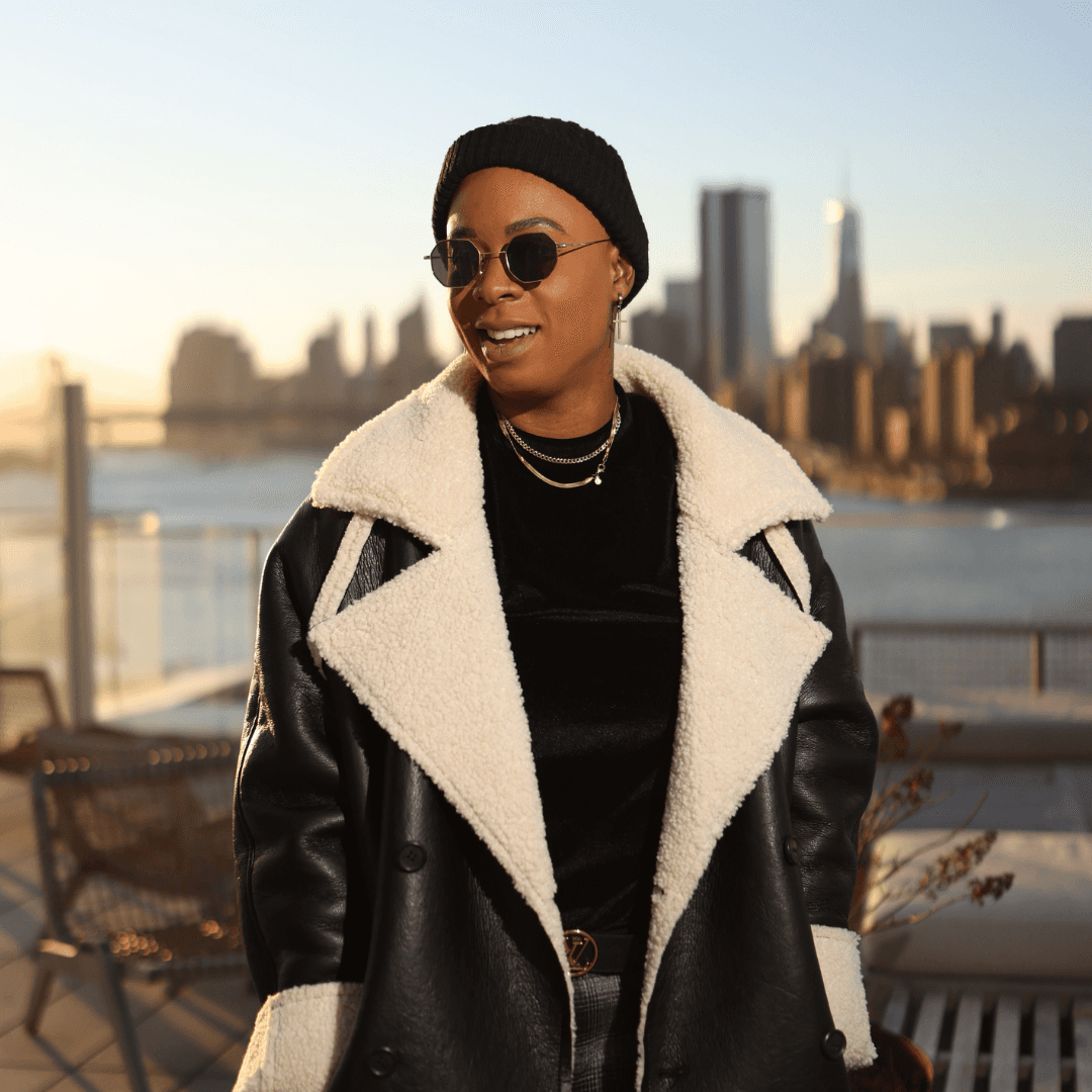 Ashleigh Warren, a person wearing a stylish black and white coat and sunglasses stands outdoors with a city skyline in the background.
